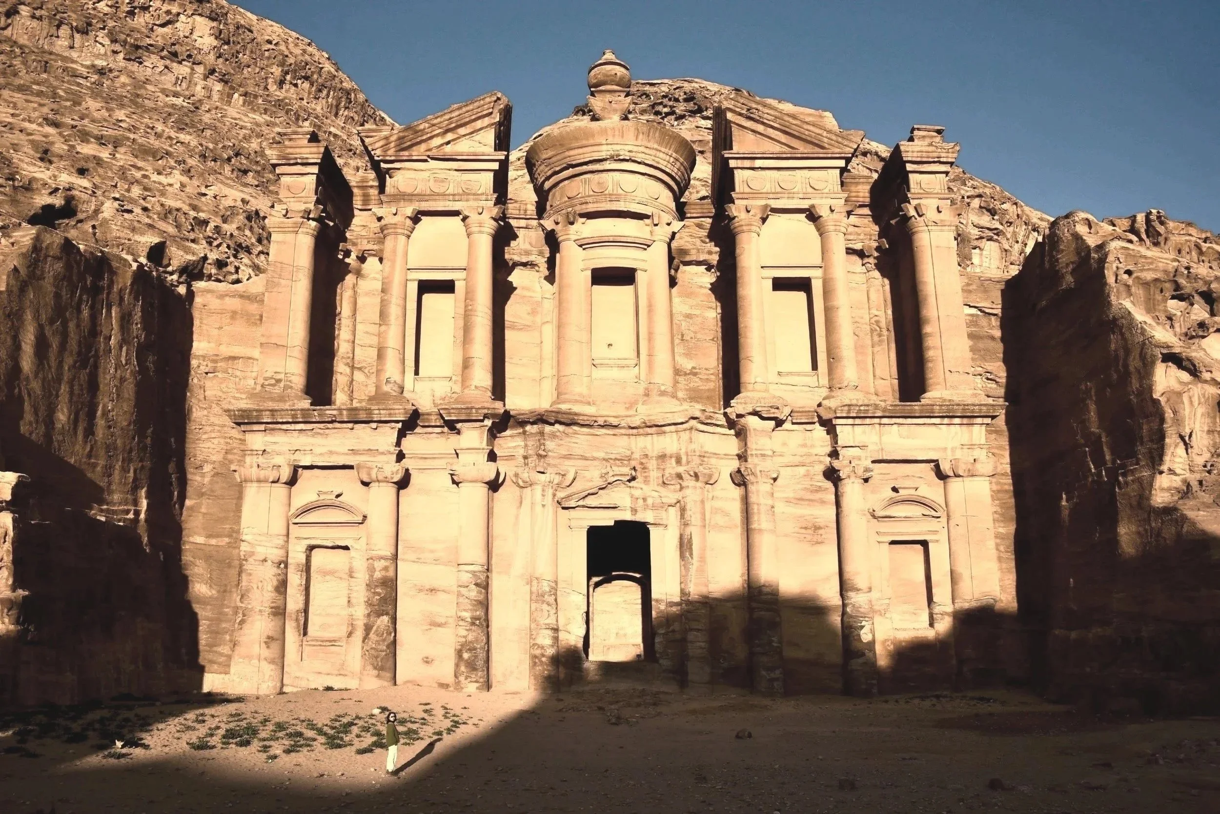 Ancient stone building with Corinthian columns and intricate details, set against rocky mountains and clear blue sky.
