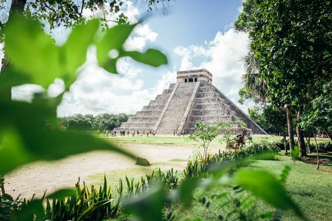 View of the ancient Mayan pyramid El Castillo at Chichen Itza, surrounded by lush green trees and grass, under a partly cloudy sky.