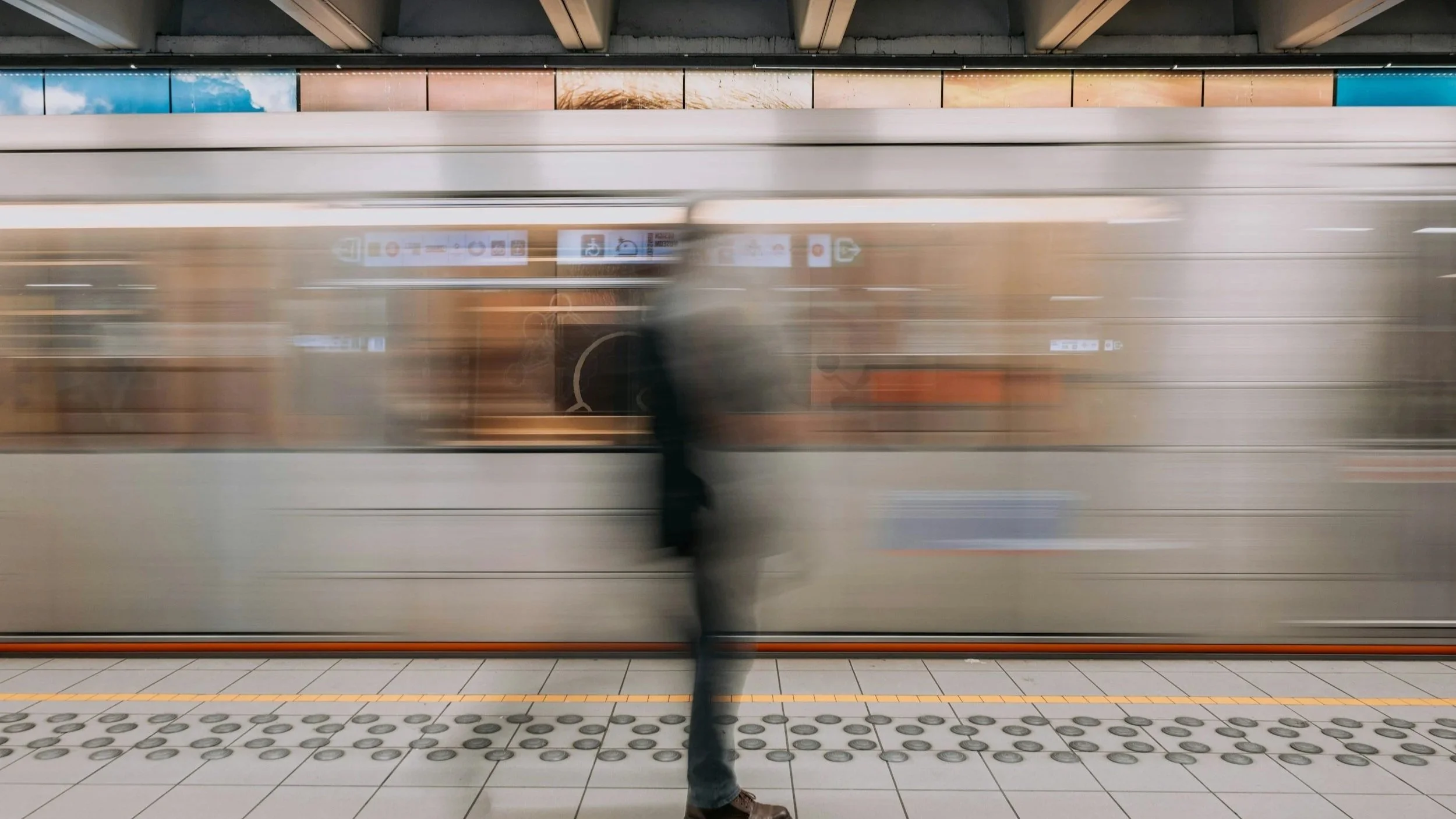 A person standing on a subway platform while a train passes by at high speed, creating a motion blur effect.