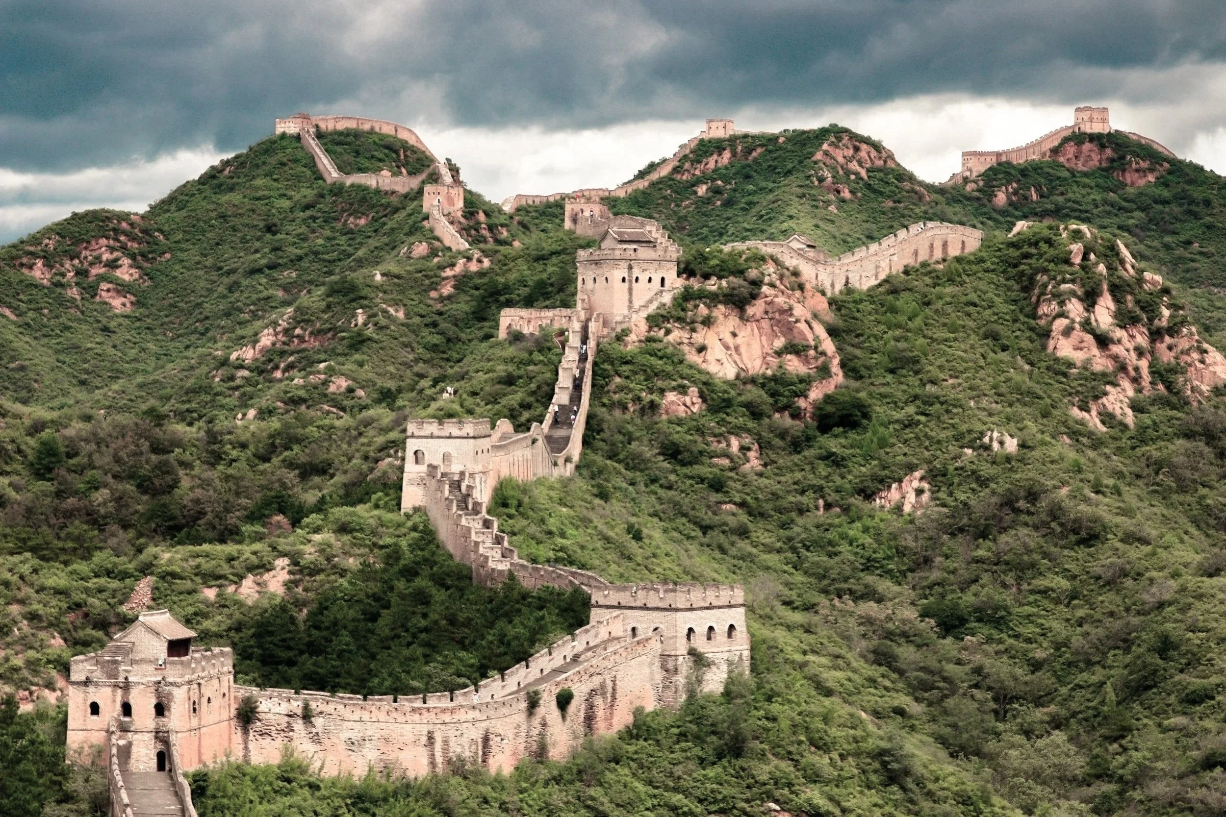 The Great Wall of China winding over green hills and mountains under a cloudy sky.
