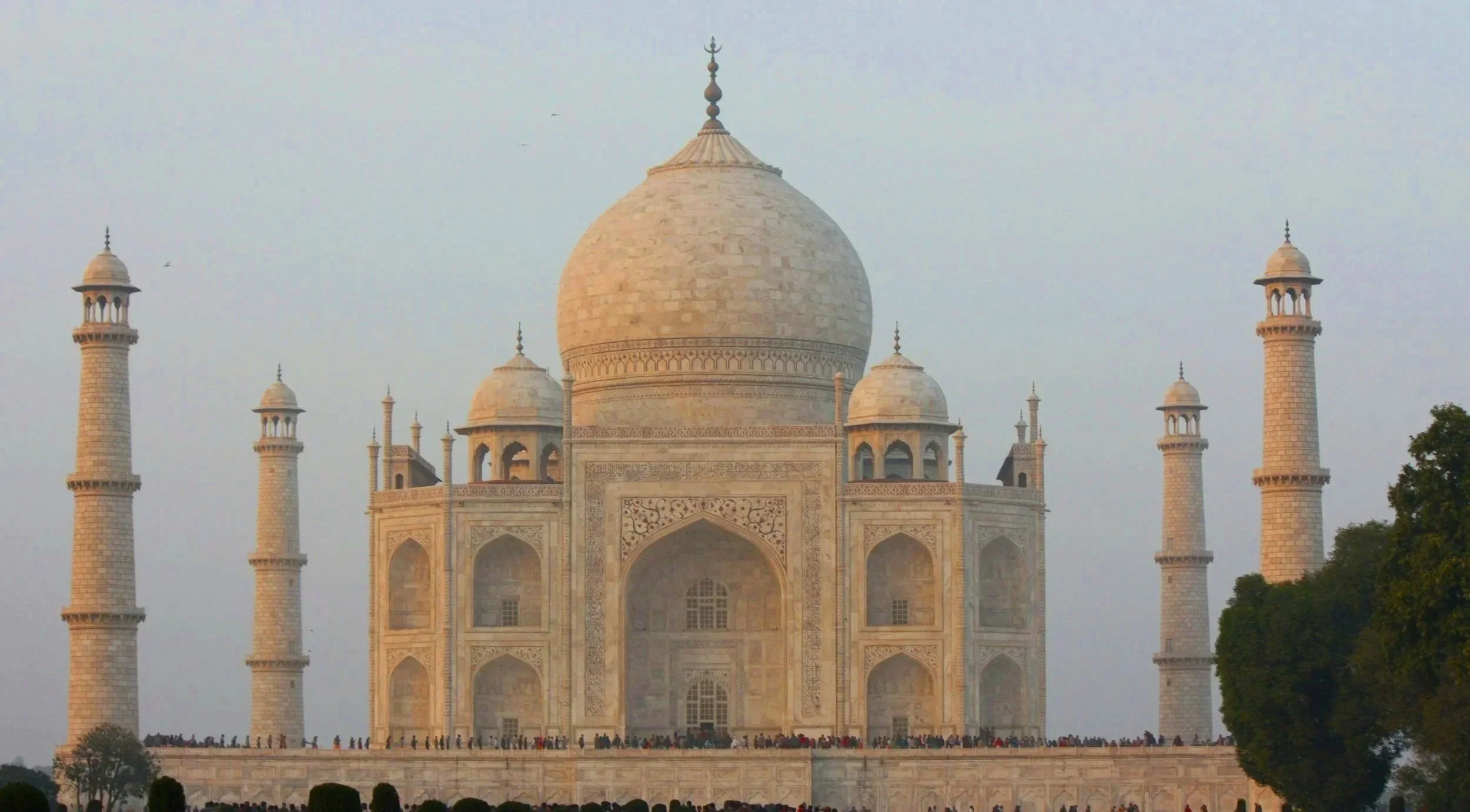 The Taj Mahal, a white marble mausoleum with a large central dome and four minarets, is shown from the front with tourists around its base.
