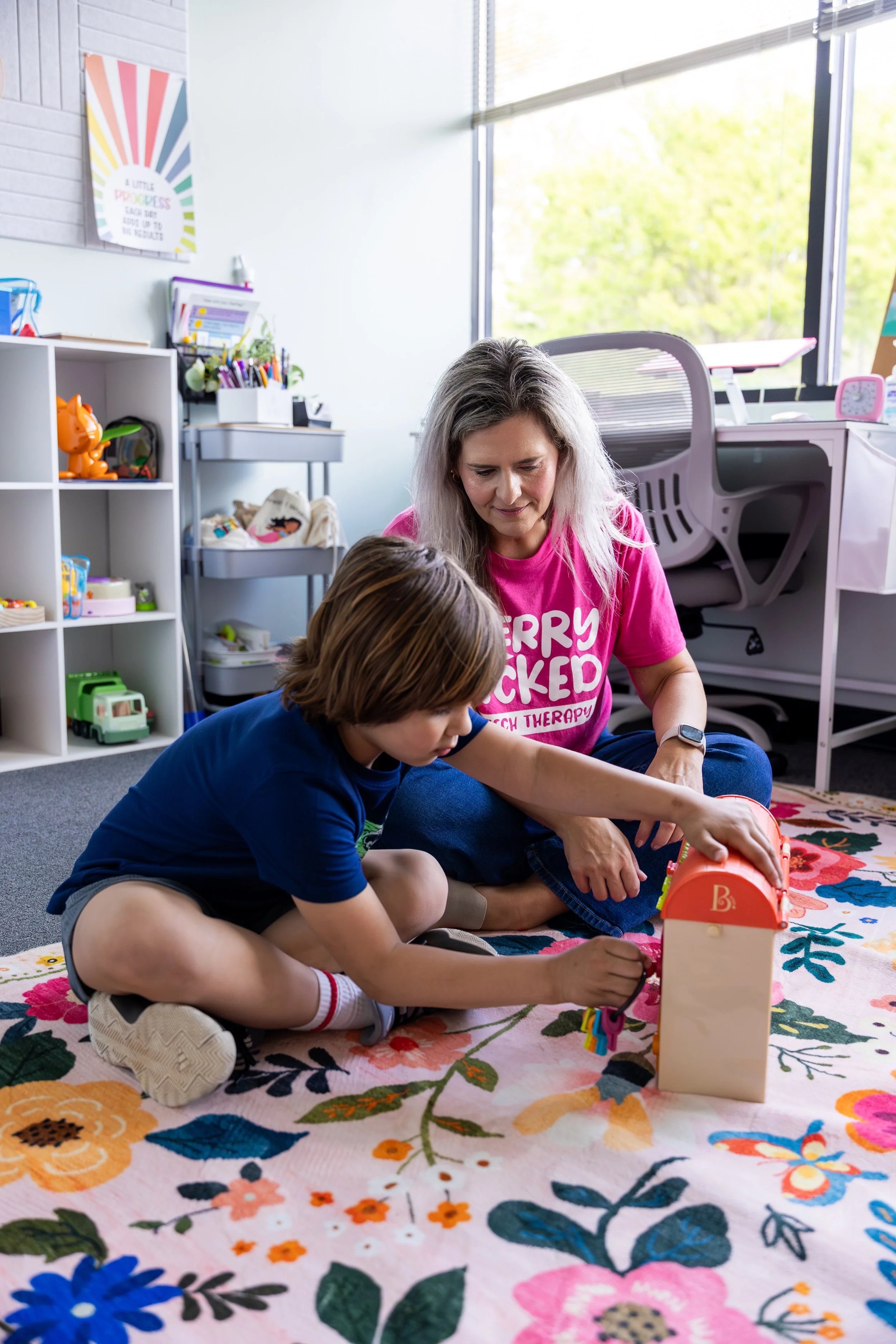 A woman and a young boy sitting on a floral rug indoors, playing with a small wooden house and colorful keys, working on improving communication skills during speech therapy for gestalt language processors in Newport News, VA.