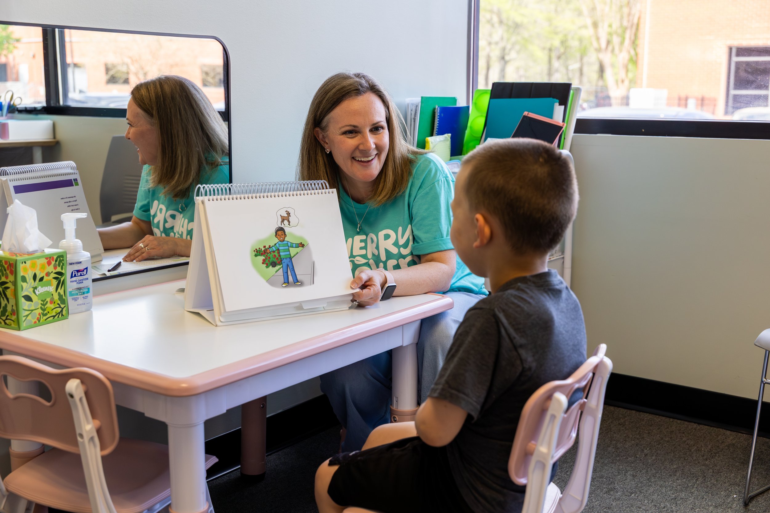 A speech therapist and a boy sitting at a table in an office, looking at pictures and smiling while working on language and vocabulary skills during speech therapy for kids in Newport News, VA.