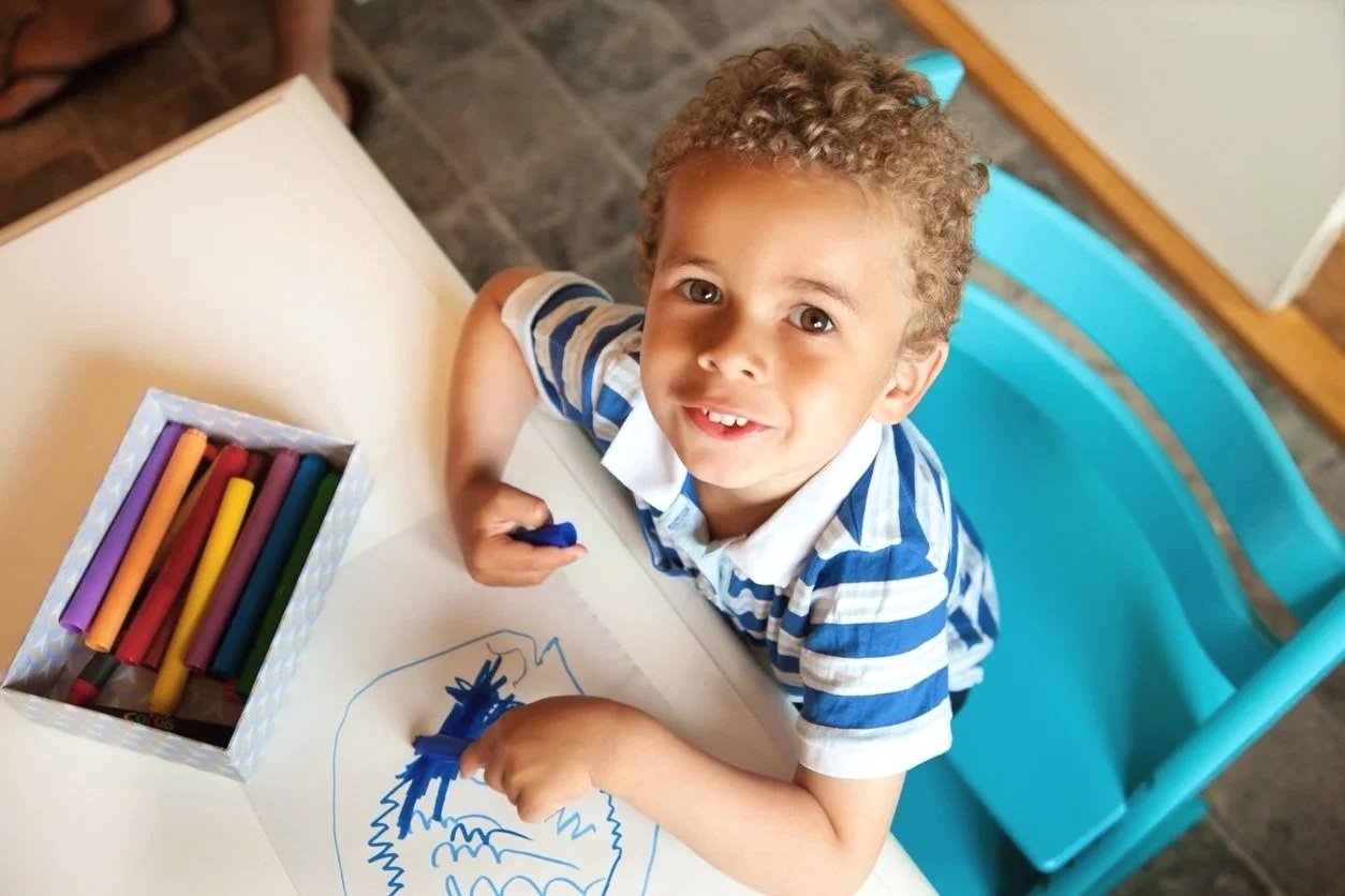 A young boy with curly hair and a striped shirt sitting at a table, drawing with colorful markers on a piece of paper. The boy is working on using his correct speech sounds while coloring during speech therapy for children in Newport News, VA.