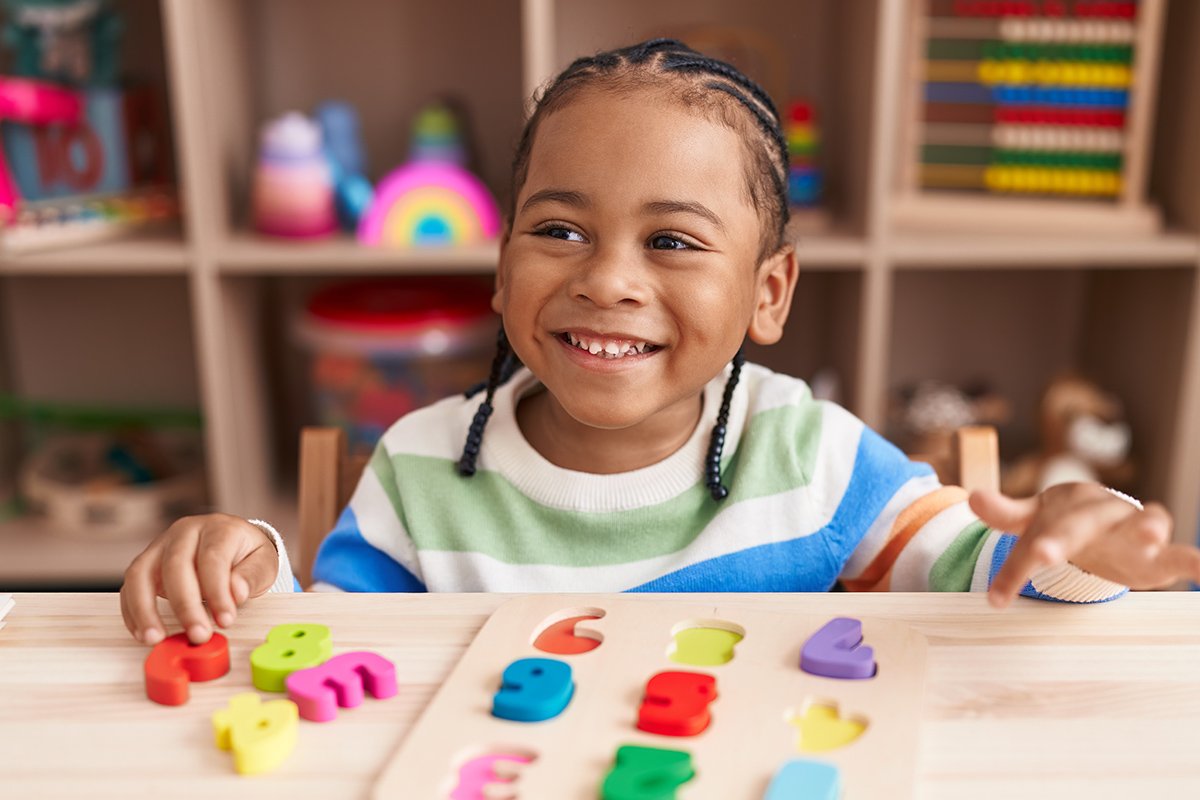 A young girl with braided hair smiling while sitting at a table with a colorful puzzle, with a bookshelf in the background. The girl is working on understanding and using language during language therapy for kids in Newport News, VA.