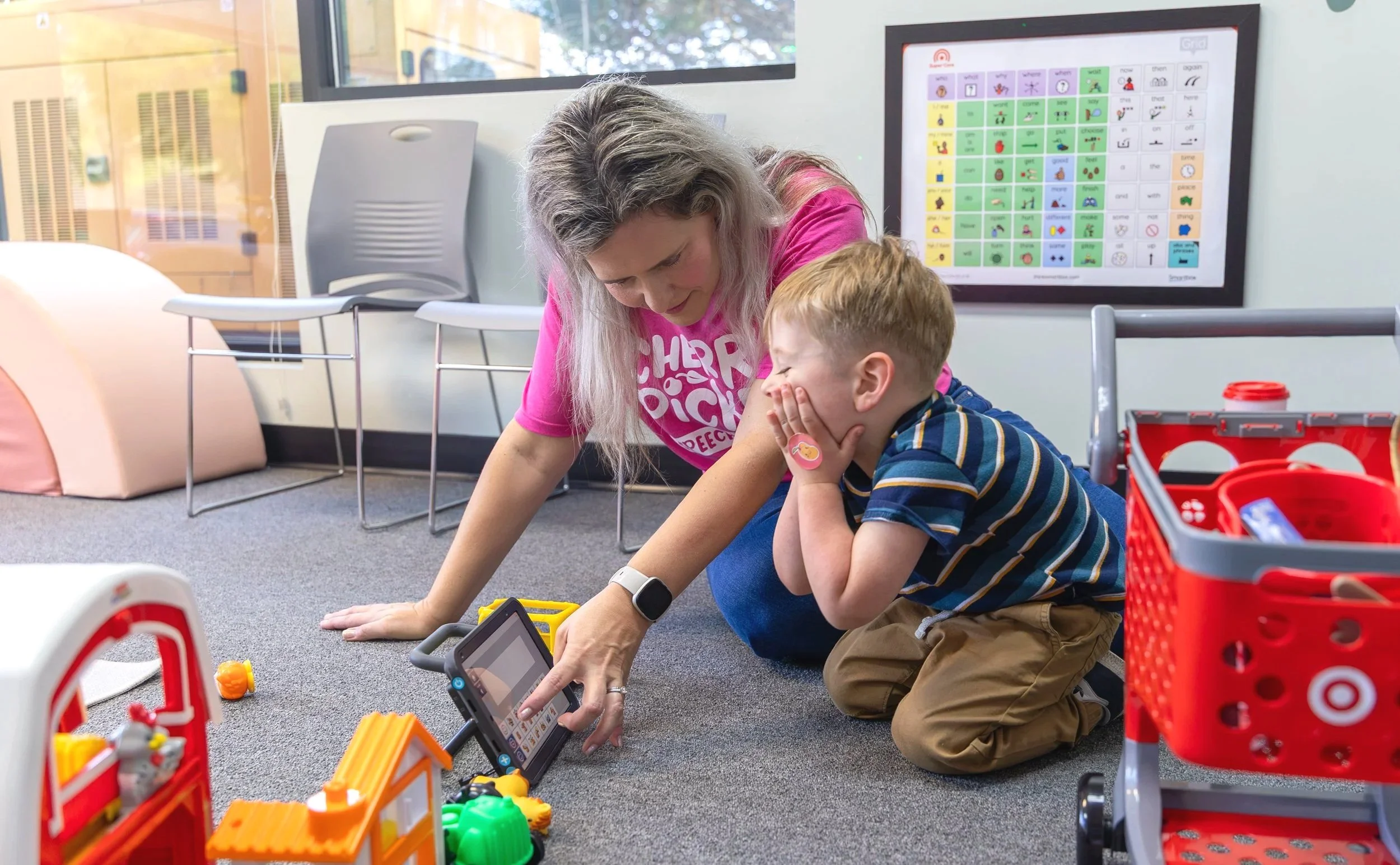 A speech therapist and a young boy are sitting on the floor, playing with colorful toys while using a tablet for communication during AAC therapy in Newport News, VA.