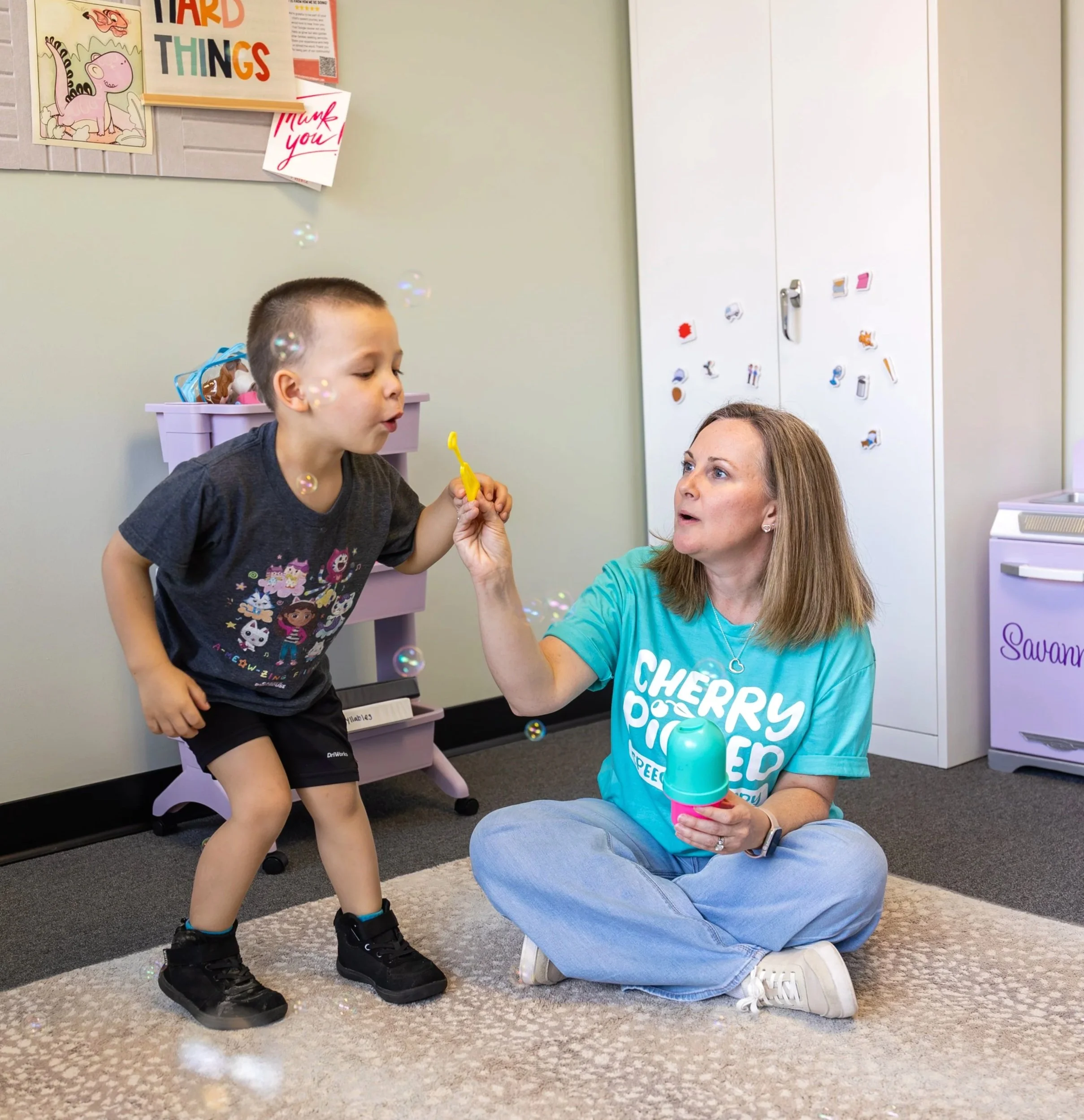 A young boy standing and blowing bubbles towards speech therapist who is sitting on the floor holding the bubble wand. The boy is participating in play-based, child-led speech therapy for gestalt language processors in Newport News, VA.