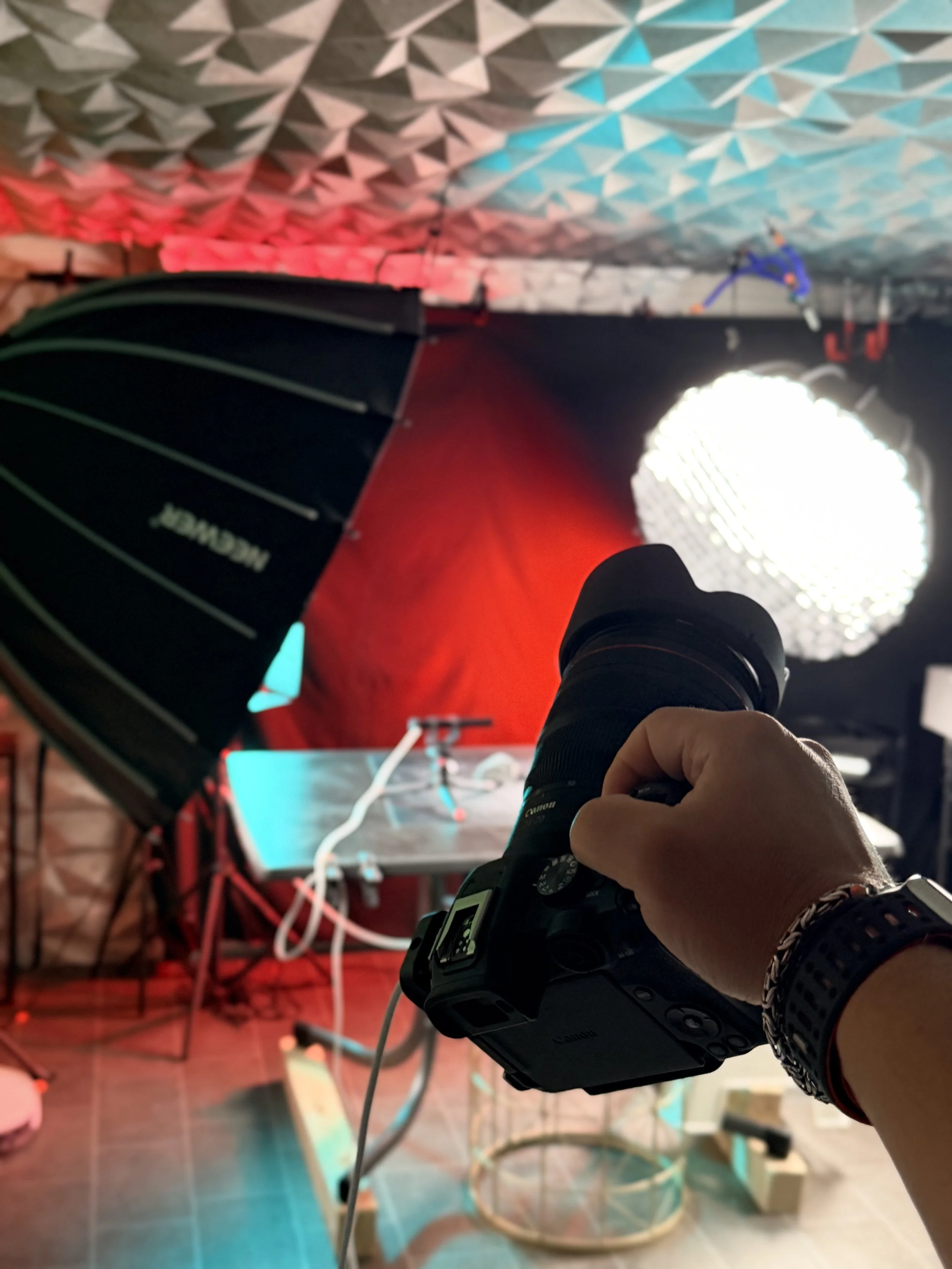 Eine Hand hält eine Kamera, im Hintergrund sind Studiobeleuchtungen und eine Arbeitsfläche mit Kabeln und technischen Geräten sichtbar.