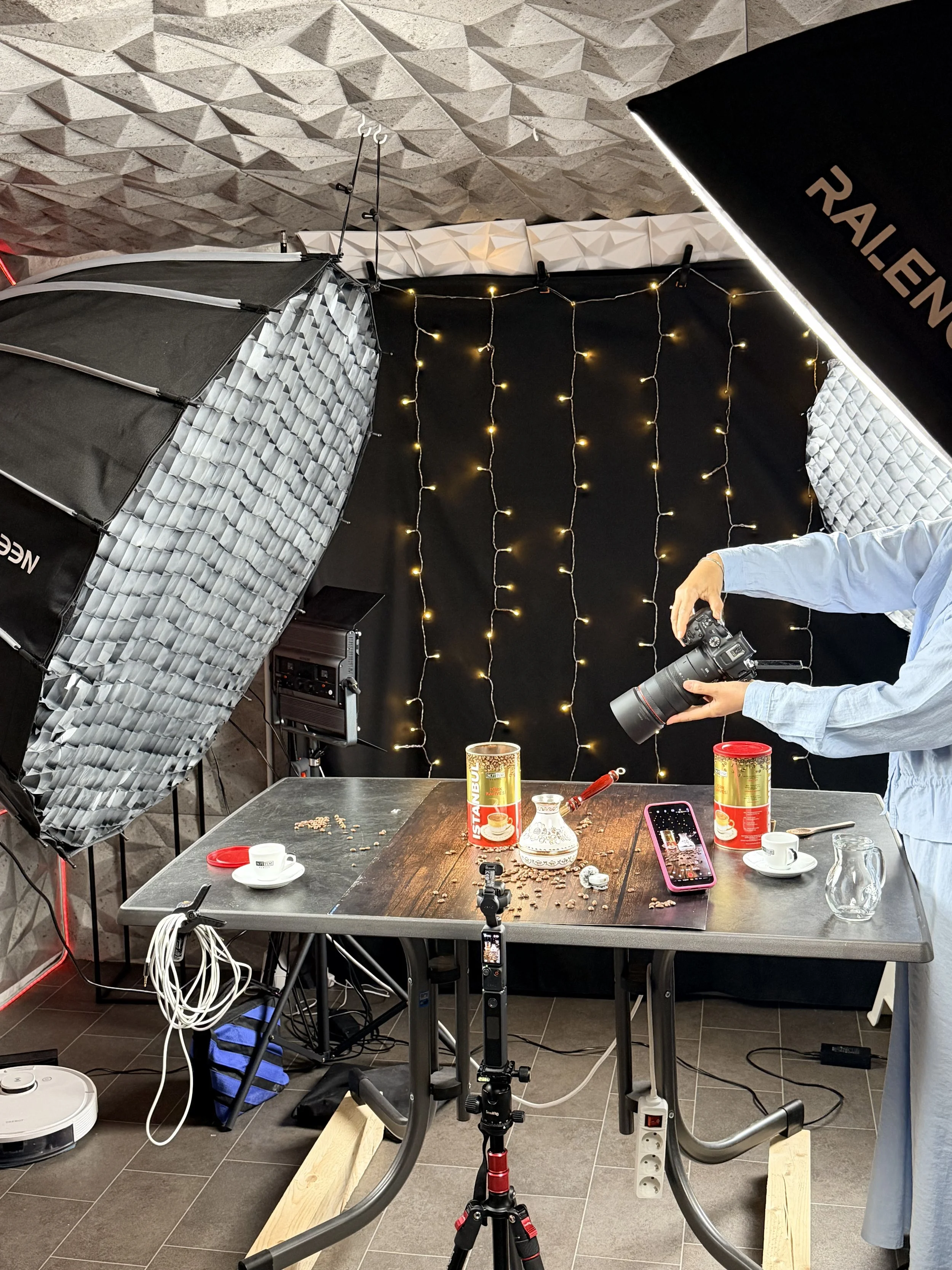 Ein Fotostudio mit Beleuchtungsausrüstung, einem Tisch mit Kaffeetassen, Keksen, einem Smartphone, einer Kamera, und verschiedenen Kabeln, im Hintergrund eine schwarze Wand mit Lichtergirlanden.