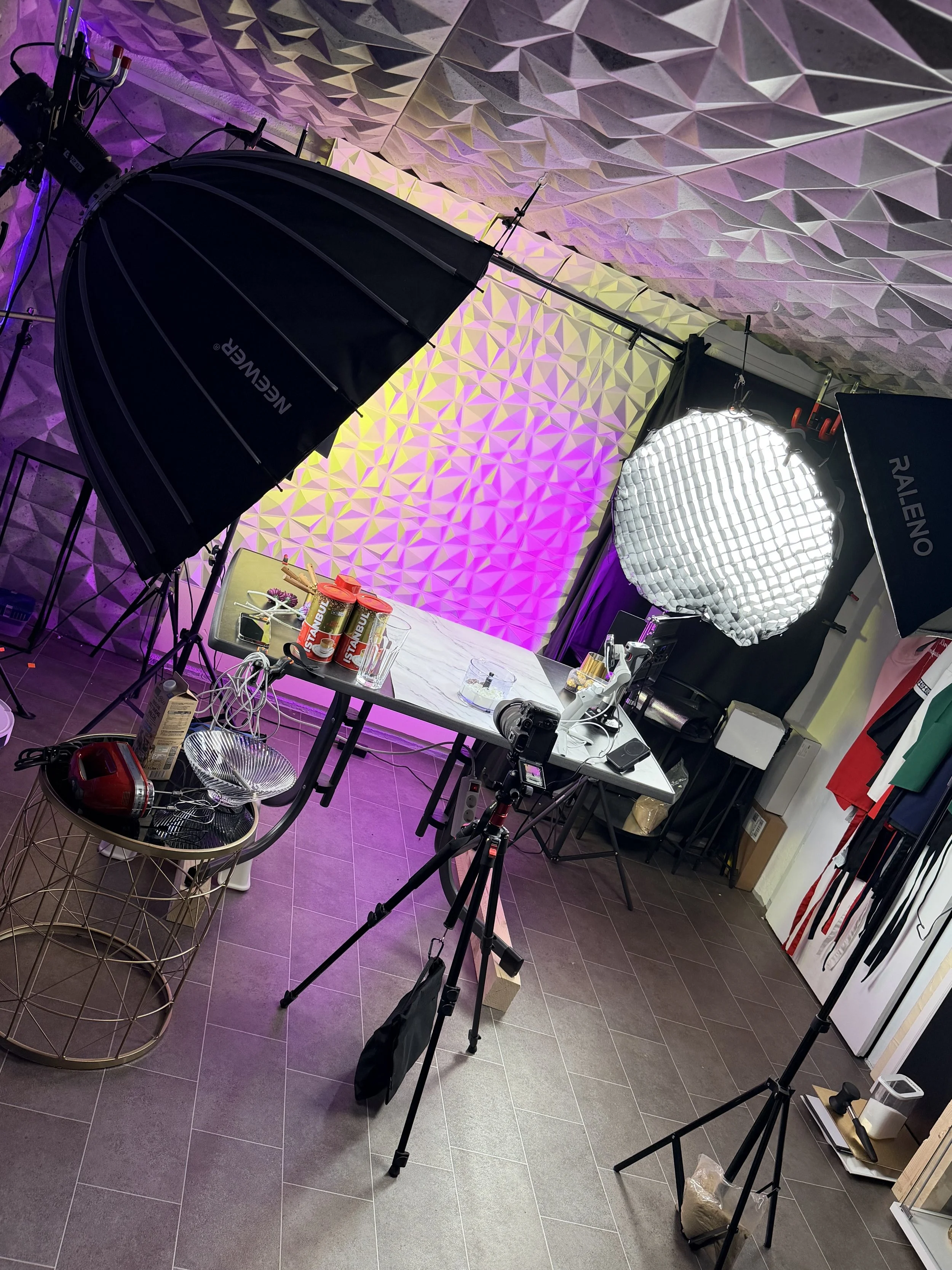 Aufnahme eines Fotostudios mit professionellem Studioequipment, Lichtquellen, einem Tisch mit verschiedenen Gegenständen und einem Hintergrund mit geometrischem Muster in lila, gelb und grau.