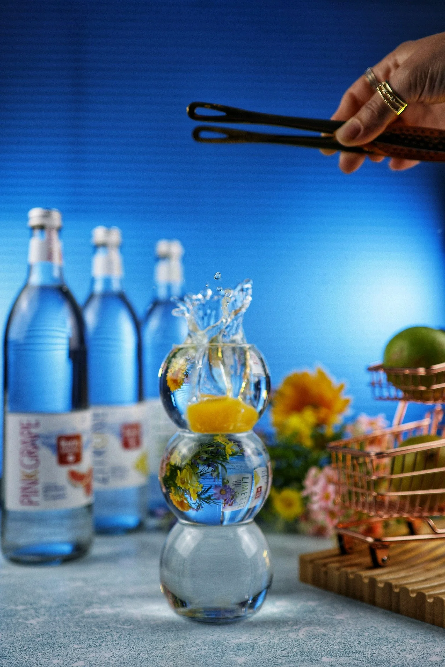 Mehrere Wasserflaschen im Hintergrund, eine kleine Vase mit Wasser, eine Zitrone und Blumen im Wasser, ein Korb mit Limetten und Blumen, ein metallener Minitisch mit Holzrost. Hintergrund ist blau.