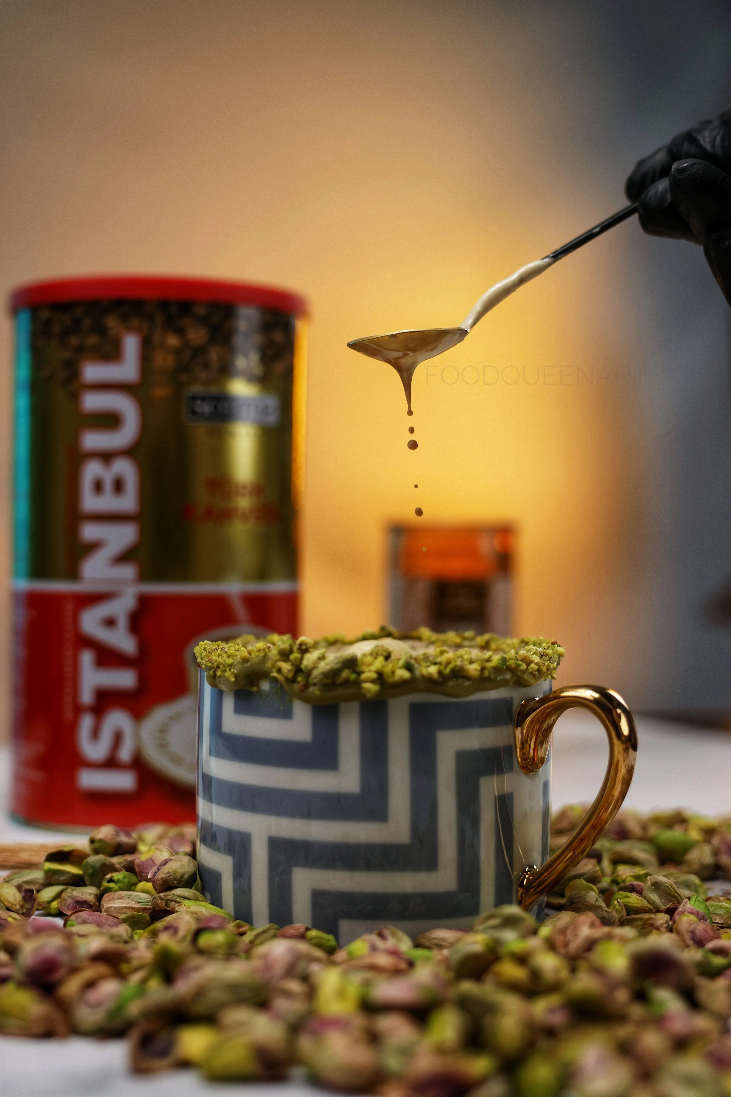 Tasse mit Pistazien und Pistazienkerne, im Hintergrund Pistaziendose und ein Löffel mit Pistaziencreme, umgeben von Pistazienkerne auf einem Tisch.