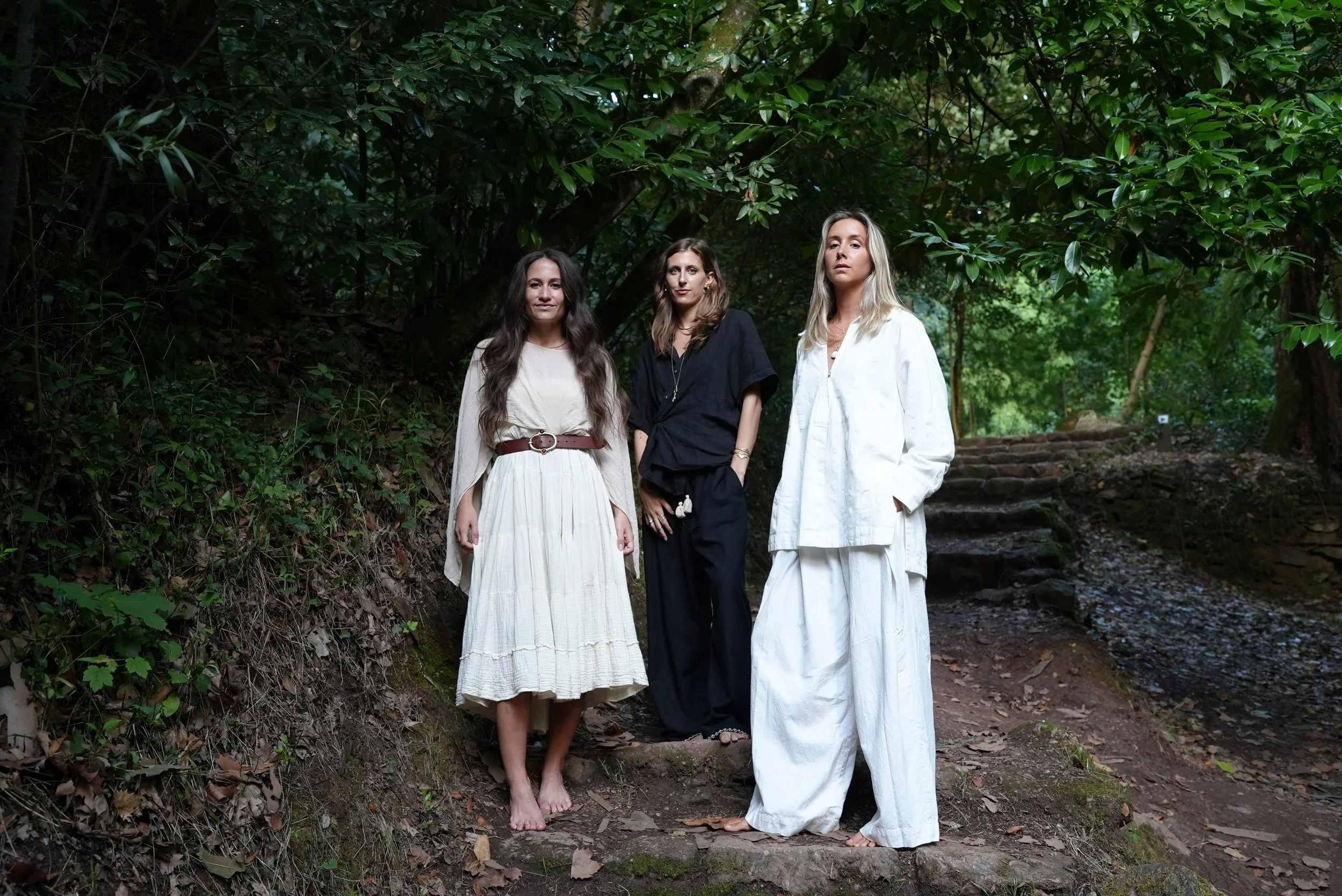 Three women standing on a forest trail, dressed in white and black flowing outfits, with trees and greenery in the background.