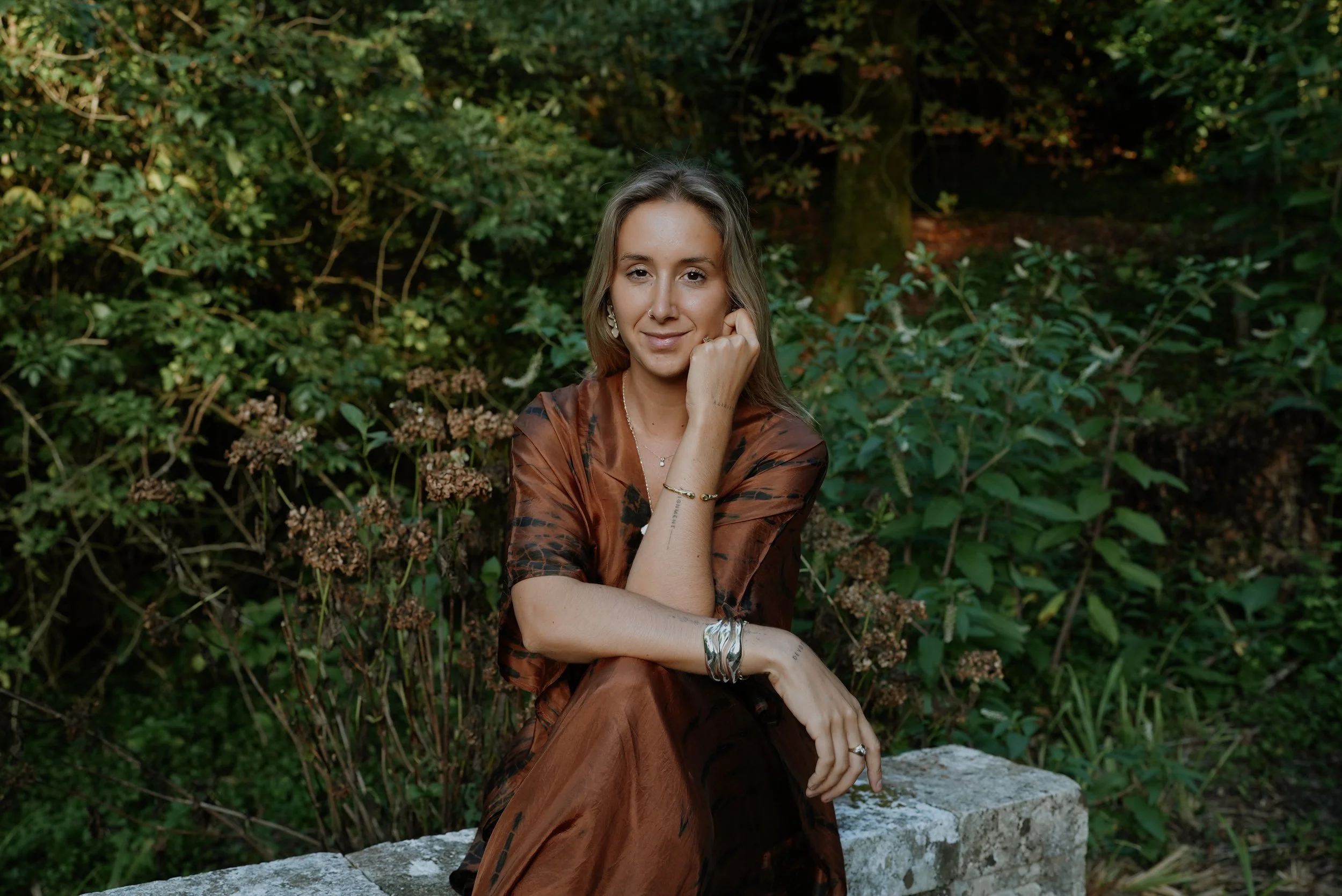 A woman sitting outdoors on a stone bench in a lush green garden.