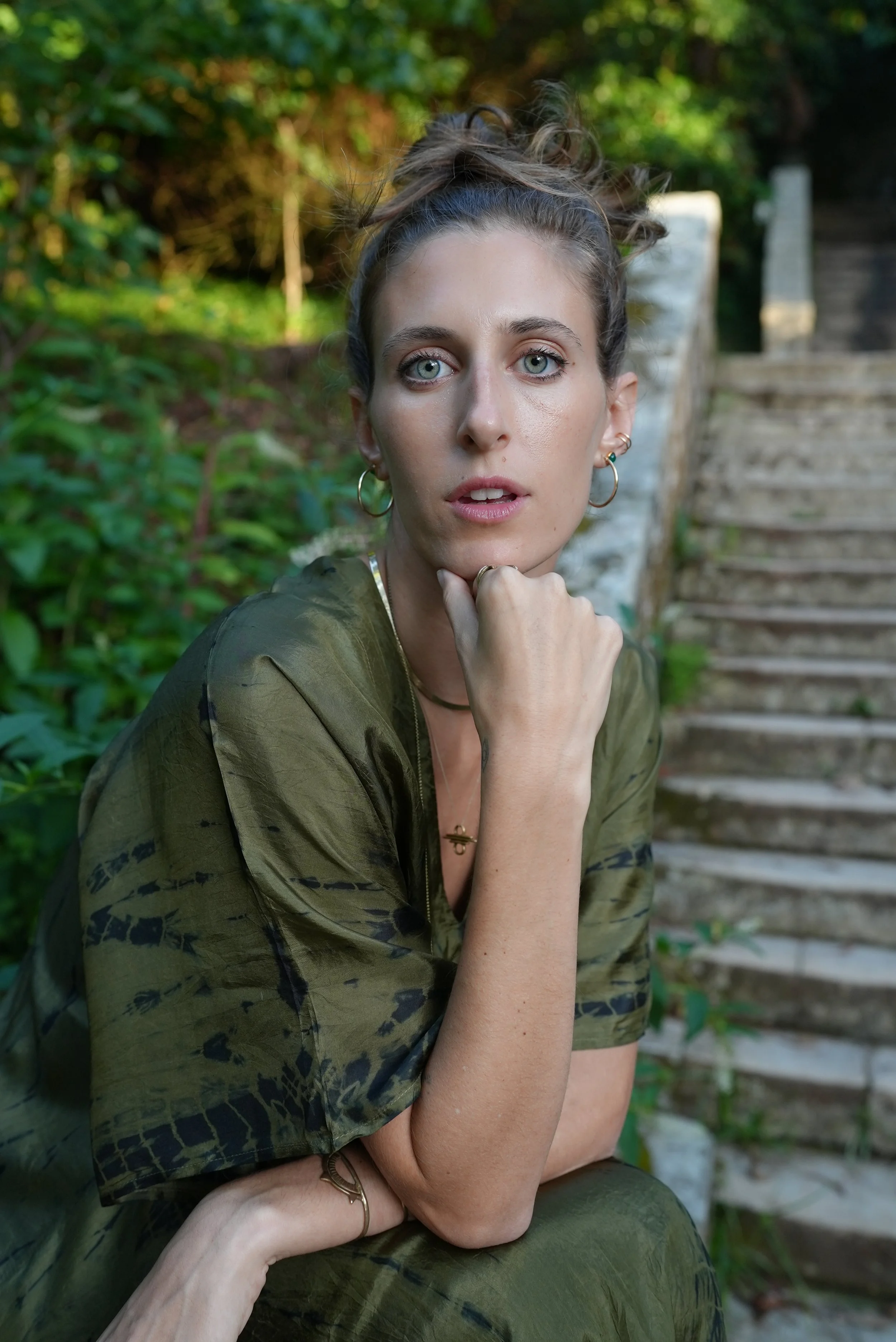 A woman with blue eyes and curly hair tied up in a bun, wearing a green patterned shirt with rolled-up sleeves, sitting outdoors on stone stairs surrounded by green foliage.