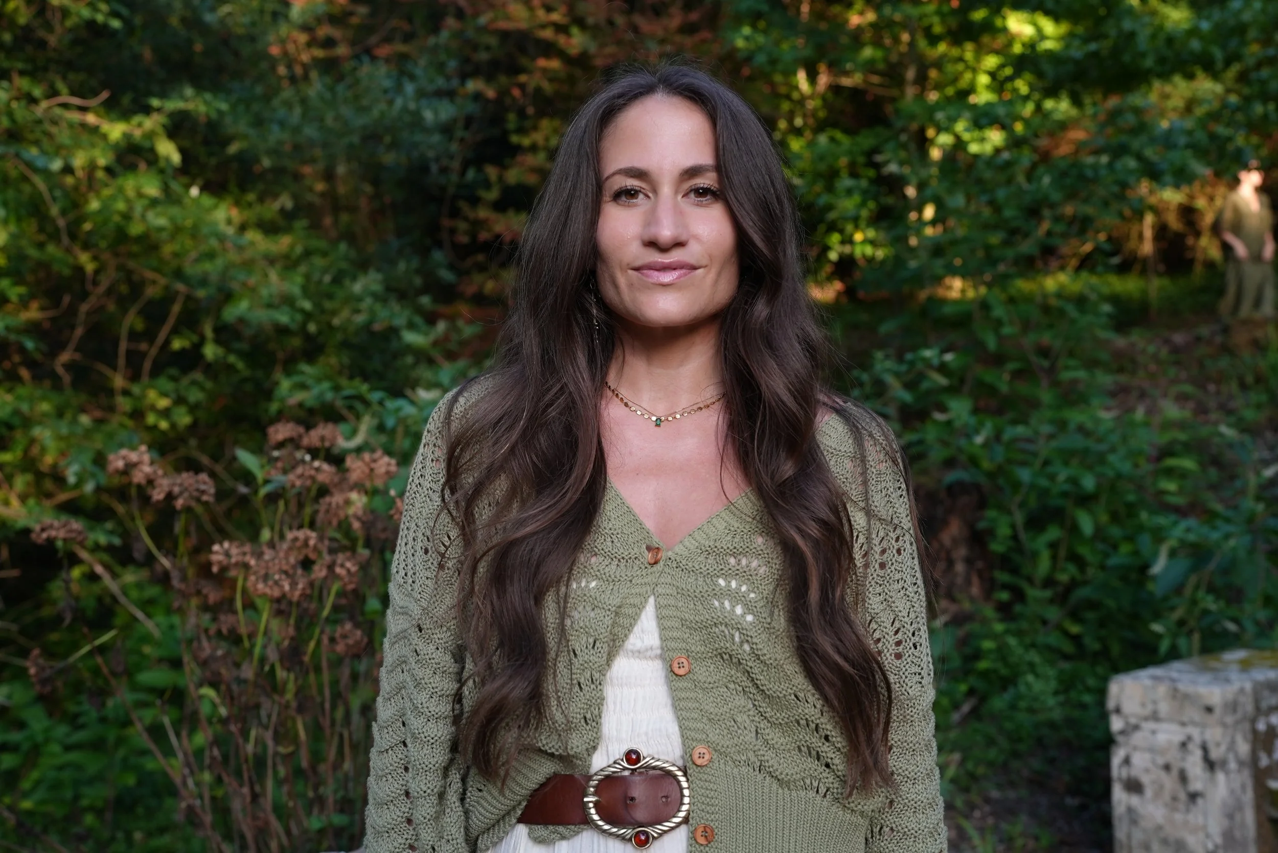 Portrait of a woman with long brown hair and a gentle smile standing outdoors in a lush green garden with trees and plants.