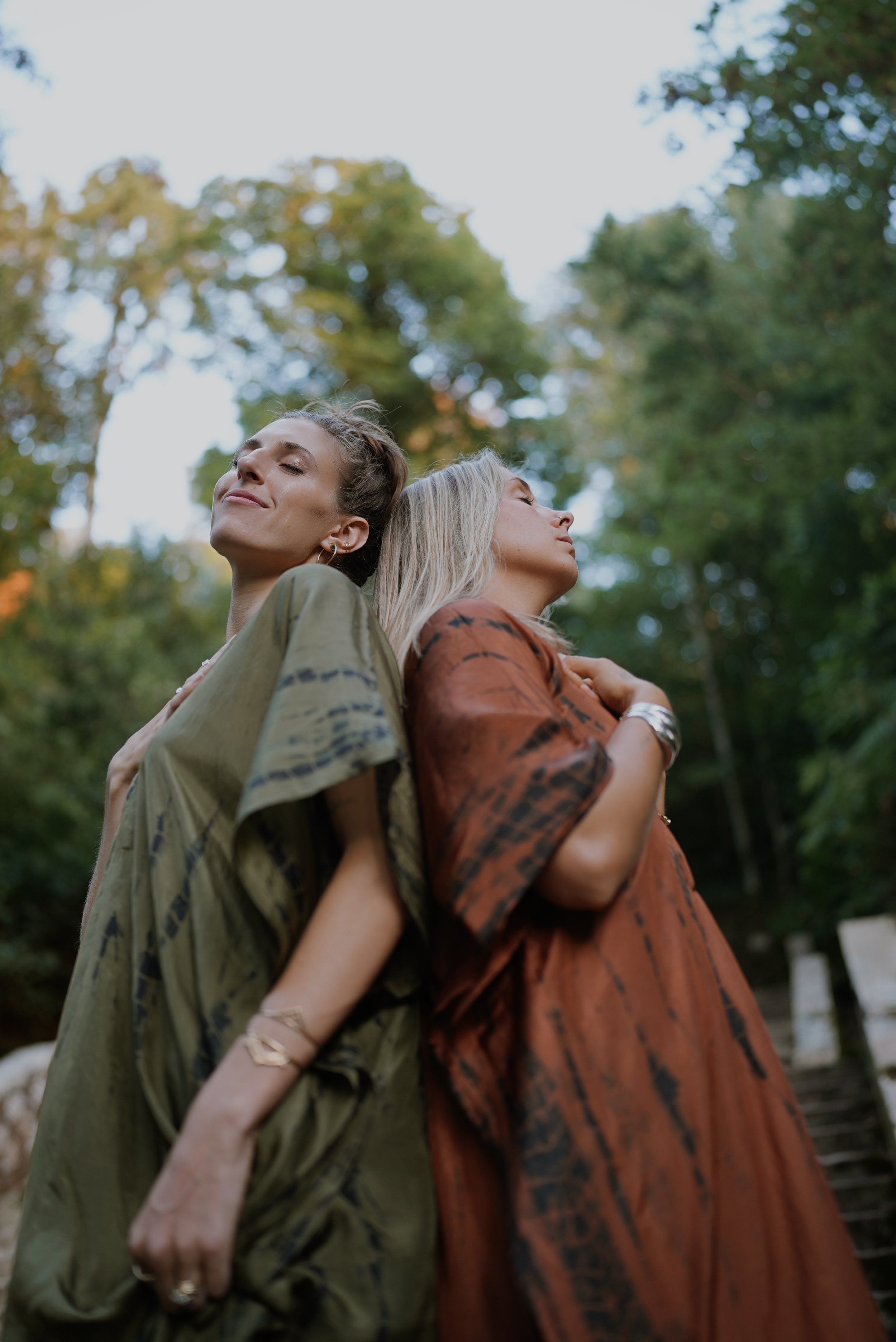 Two women standing outdoors in a park with trees in the background, their backs leaning against each other with eyes closed and serene expressions.