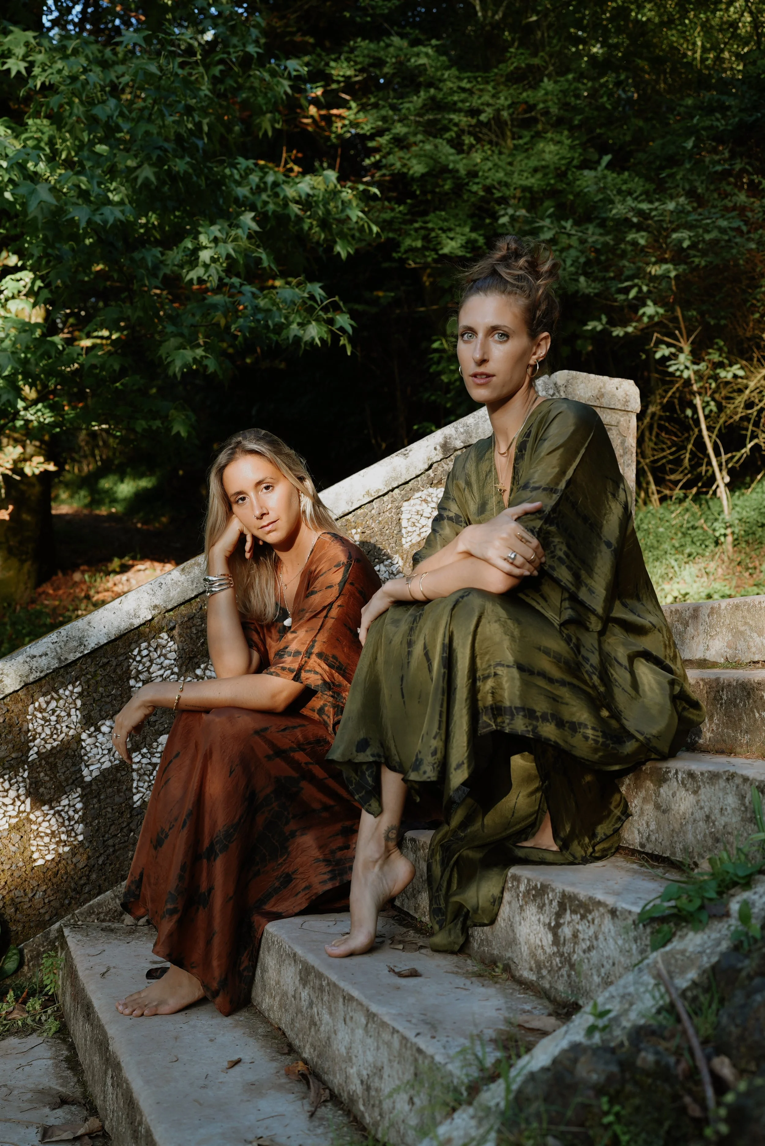 Two women sitting on stone steps outdoors, surrounded by greenery, wearing long, flowing dresses, one barefoot, with relaxed expressions.