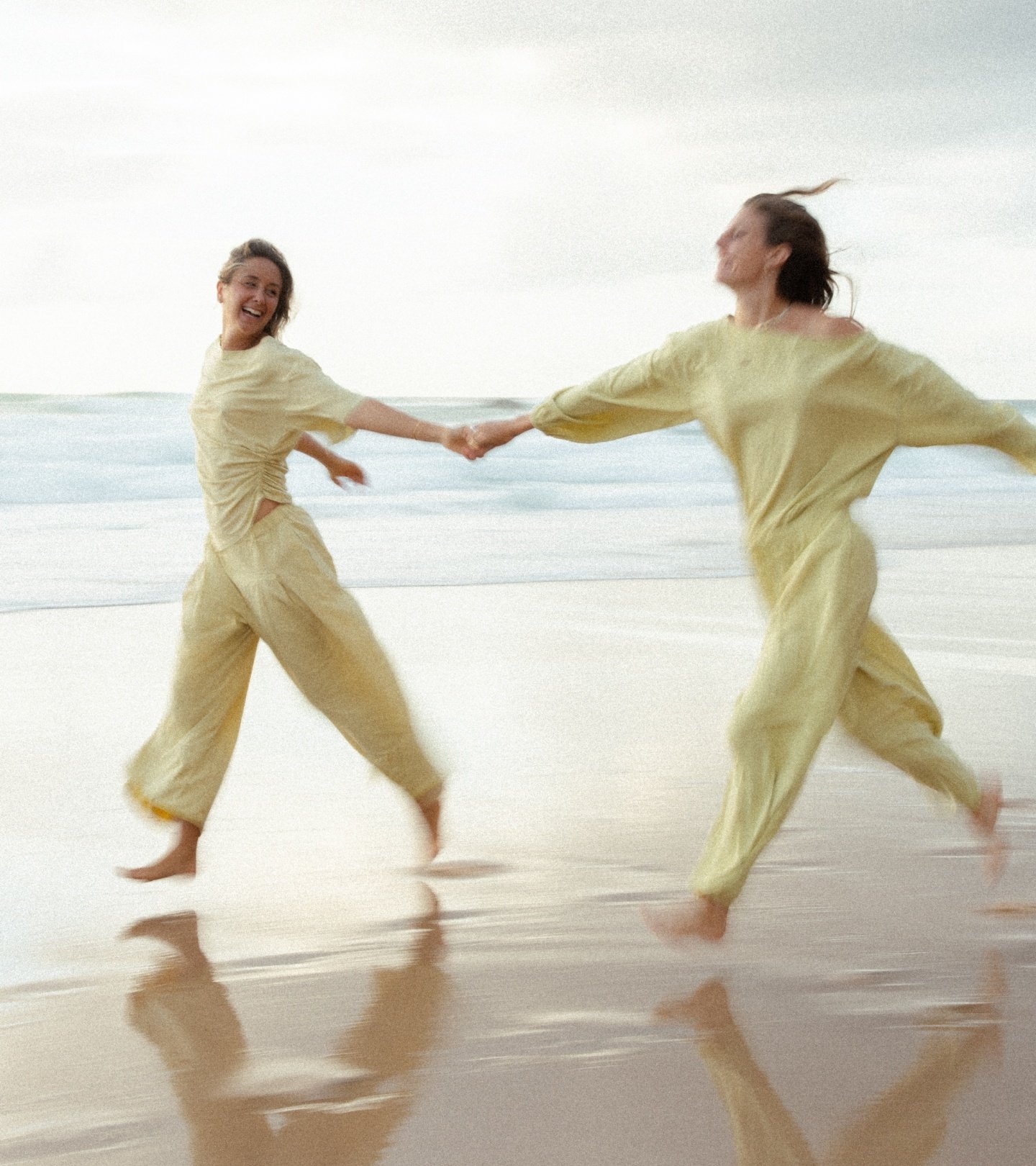 Two women dancing on a beach holding hands, wearing matching beige outfits, with ocean waves in the background.
