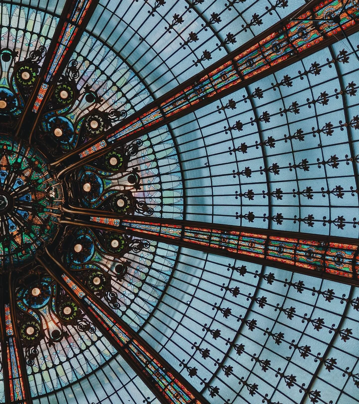 Inside view of a stained glass dome ceiling with ornate black metal framework and colorful glass panels arranged in geometric patterns.