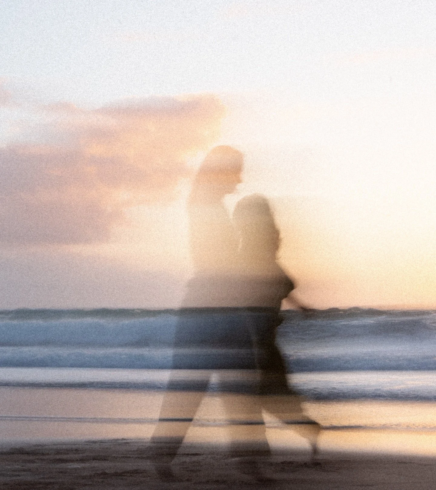 Silhouettes of a two blurry people walking along the beach during sunset with clouds in the sky.