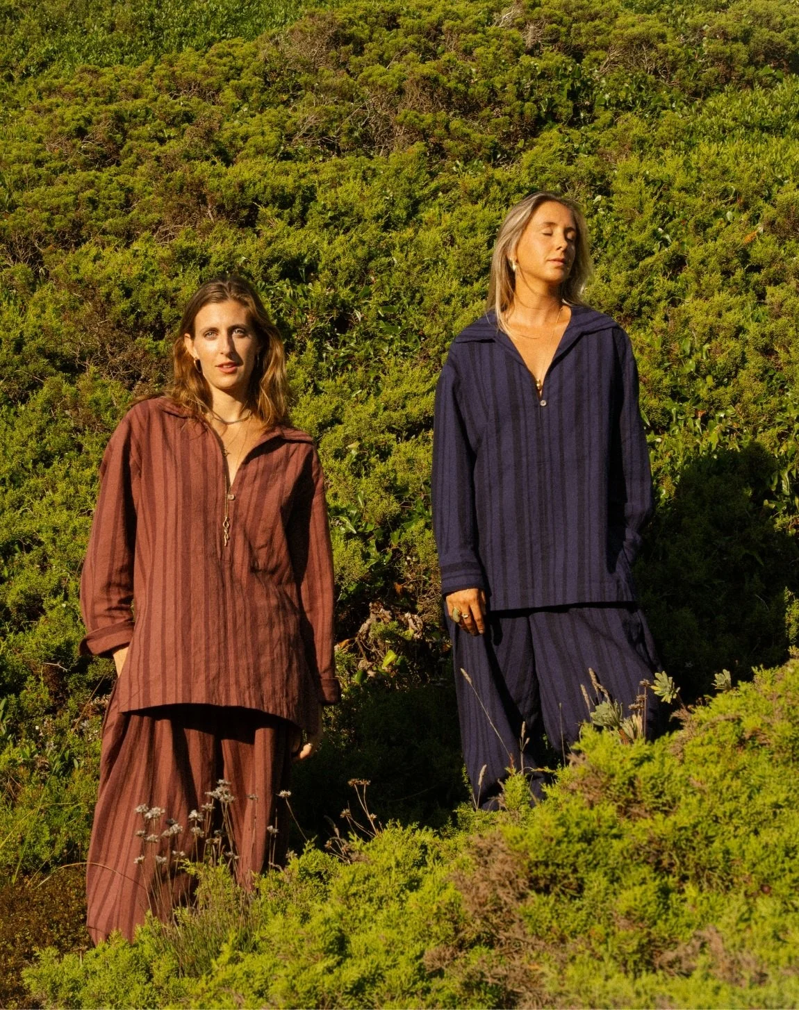 Two women standing outdoors in a lush green landscape with bushes and small plants, wearing loose, striped, long-sleeved shirts and matching pants, with their eyes closed or looking away, in warm sunlight.