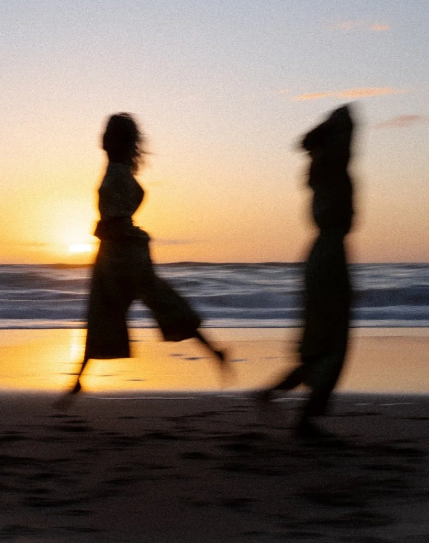 Silhouettes of two people walking on the beach at sunset, with the ocean and sky in the background.