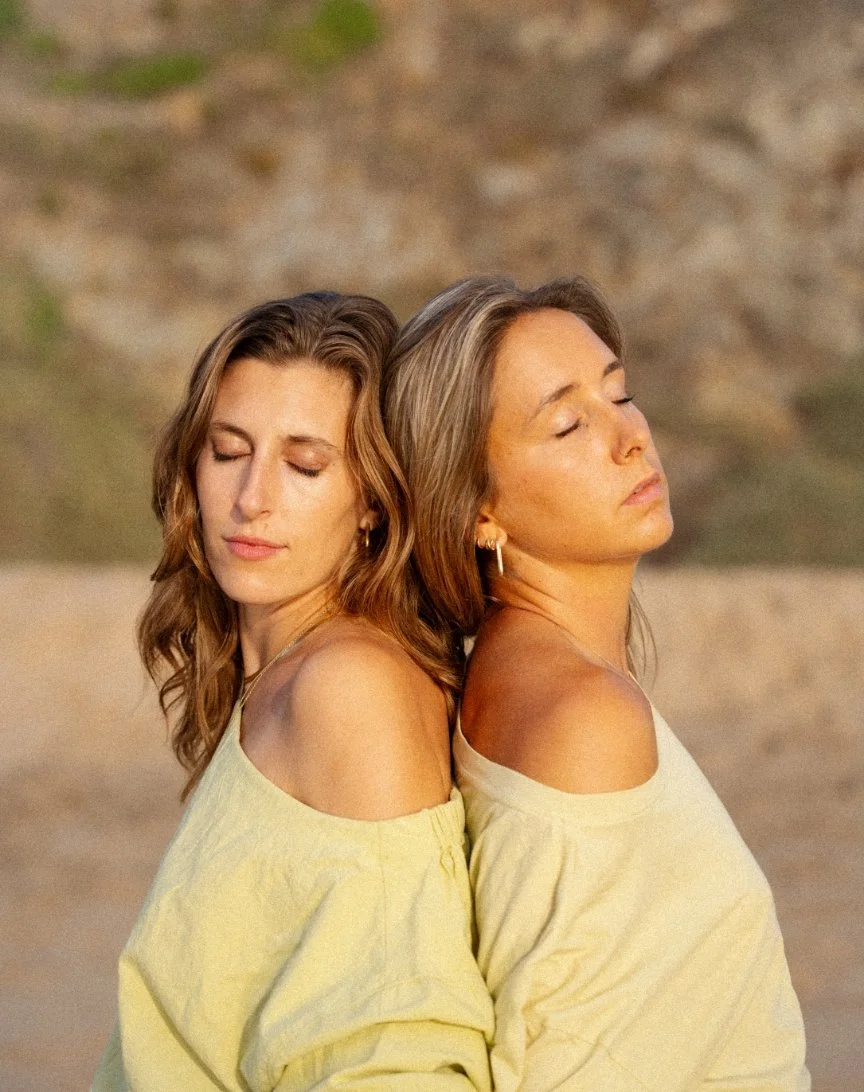 Two women standing back to back with their eyes closed, wearing off-the-shoulder yellow tops, outdoors with a blurry background.