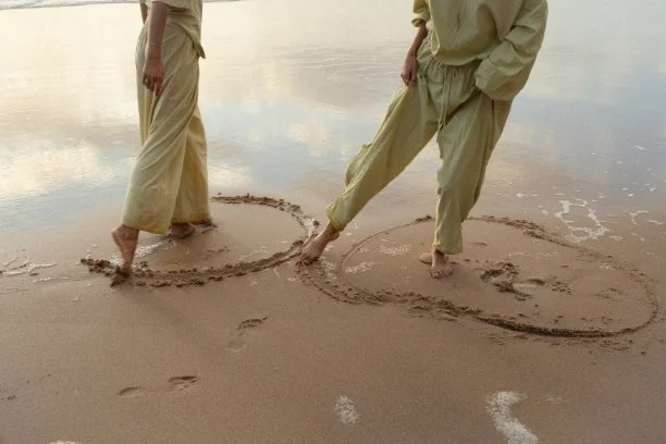 Two people standing on a beach, drawing hearts in the sand with their feet.