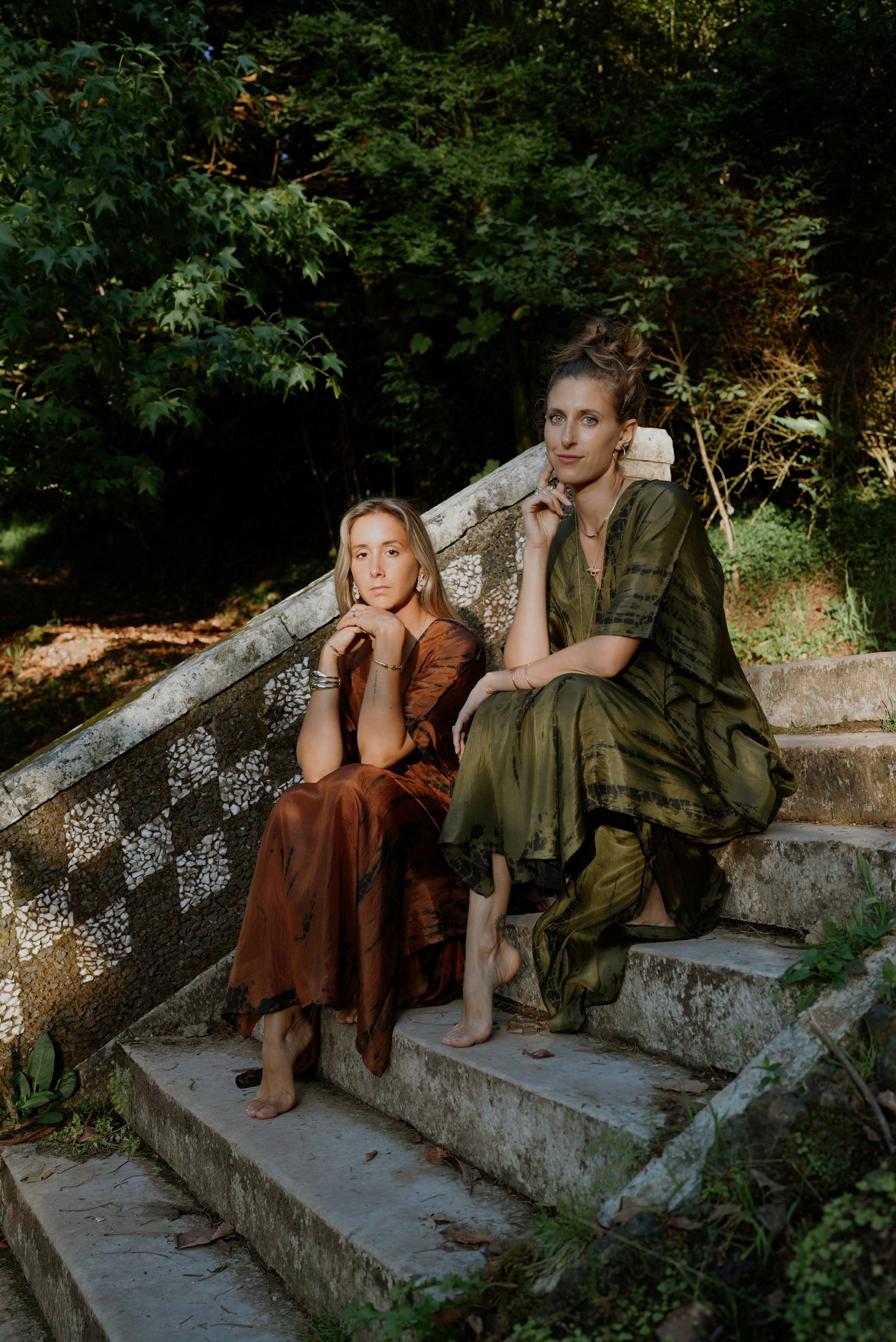 Two women sitting barefoot on outdoor stone steps in a natural, wooded setting. One woman has blonde hair and wears a rust-colored dress, resting her chin on her hand. The other has curly hair styled up and wears an olive-green dress, resting her chin on her hand. Both women have relaxed and contemplative expressions.