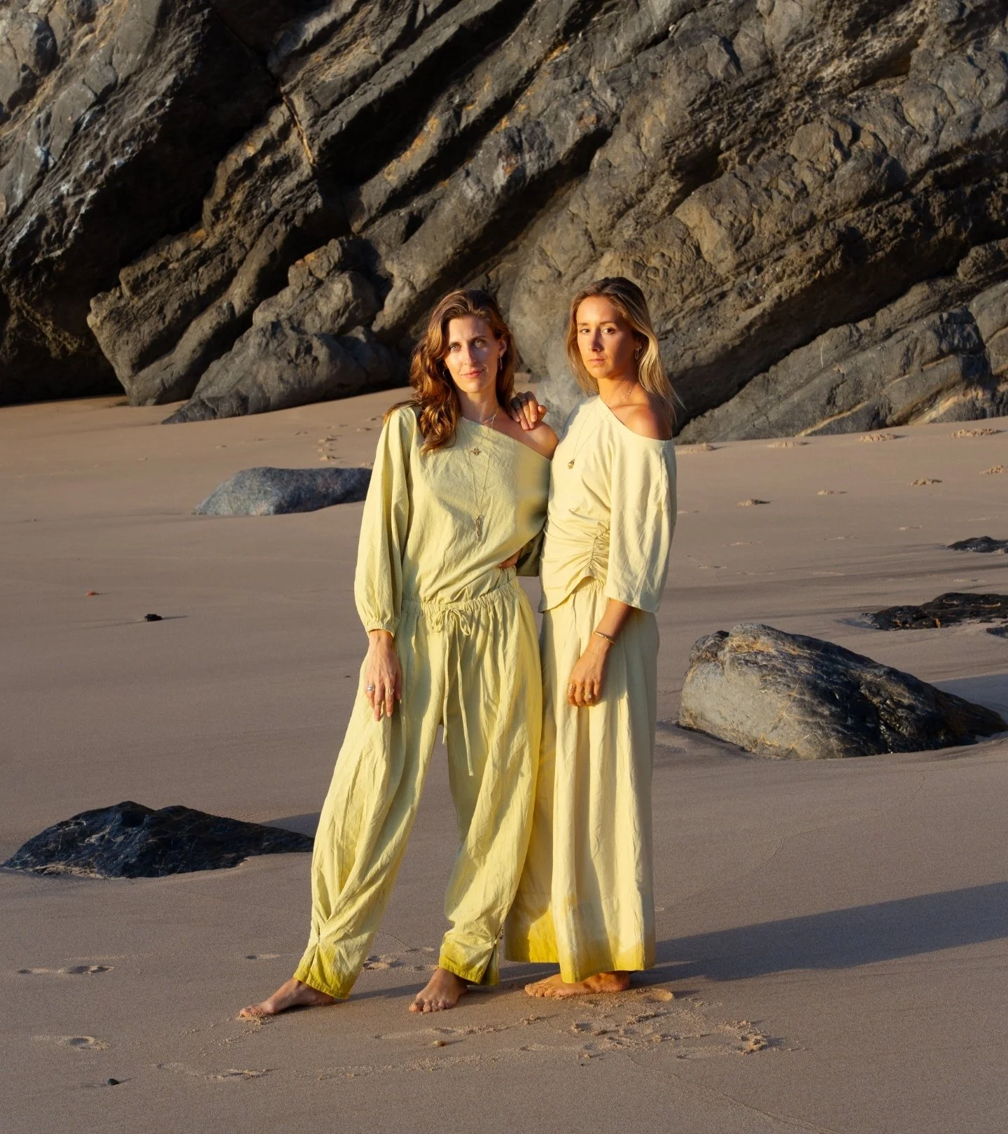 Two women standing barefoot on the beach with a large rock formation behind them, wearing matching yellow outfits and looking at the camera during sunset.