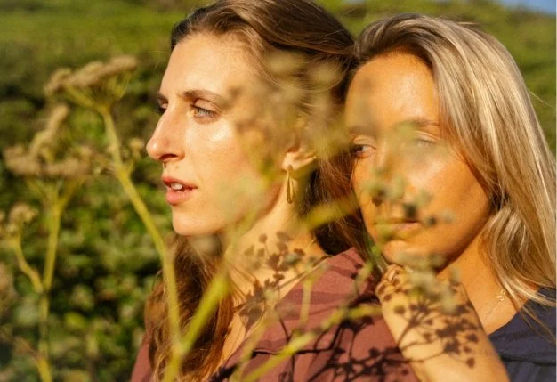 Two women sitting outdoors among plants, with a blurred background of green foliage, looking thoughtfully into the distance.