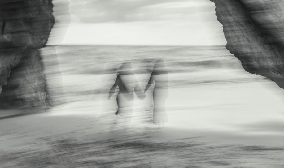 Blurry black and white photo of two people holding hands walking on a beach between rocky cliffs.