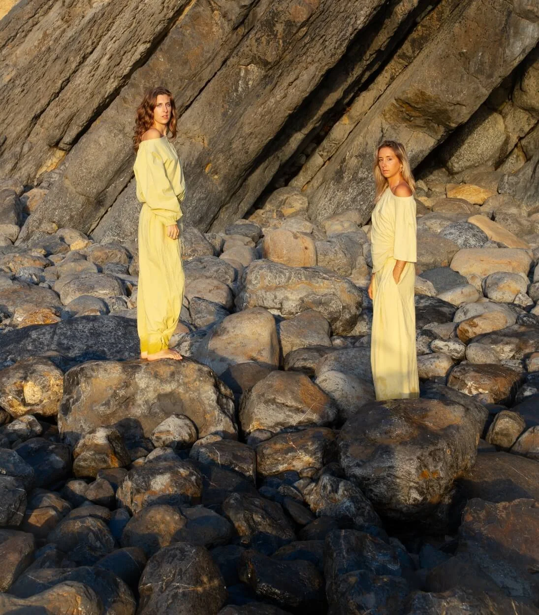 Two women in yellow outfits standing on large rocks in front of a rocky cliff with large stones around them.