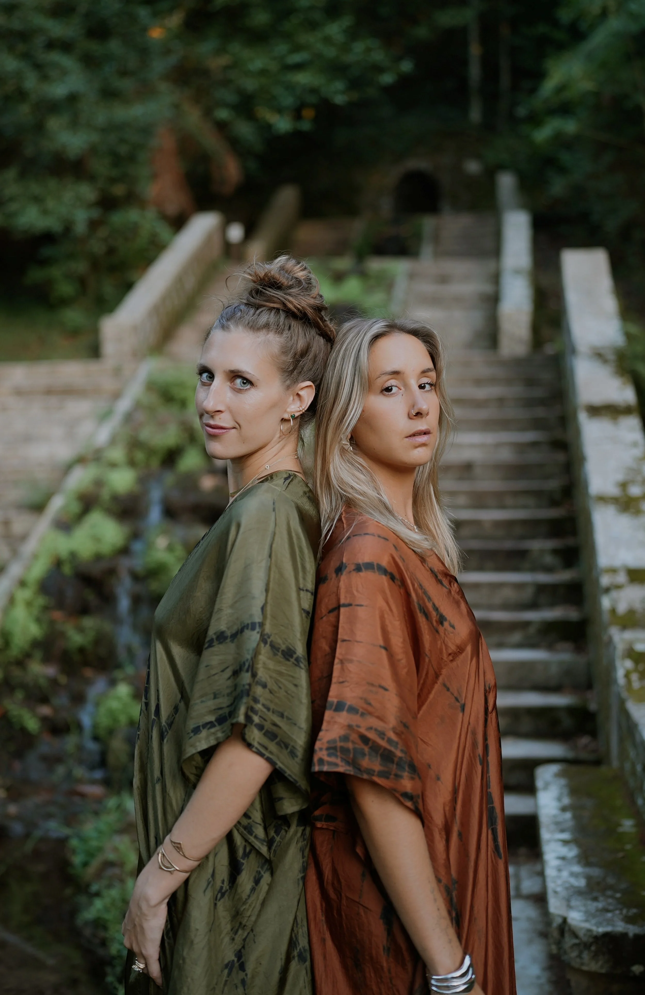 Two women standing back-to-back outdoors near a stone staircase and greenery, looking at the camera.