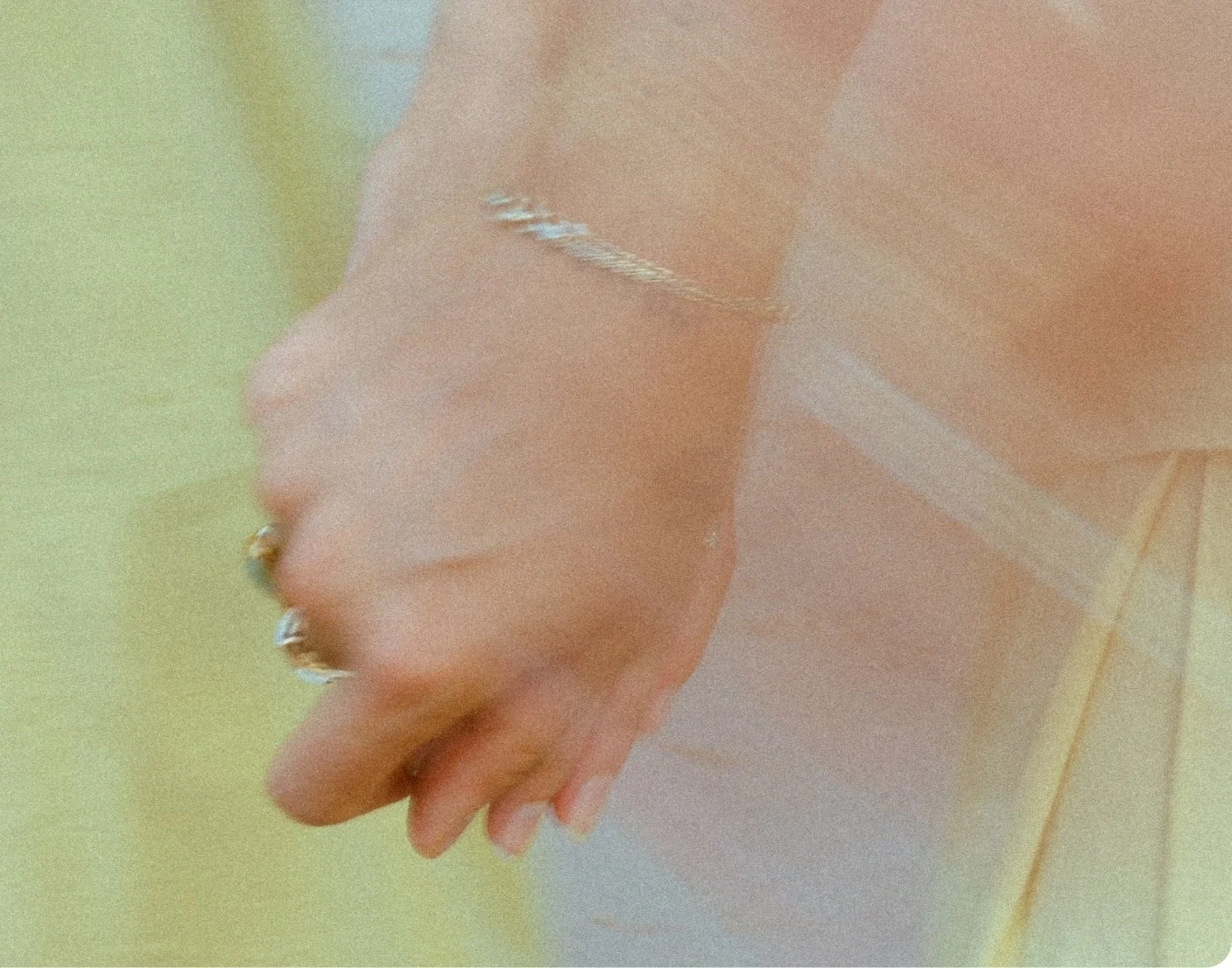 Close-up of a person's hand with rings, wearing a jewelry chain, and a white band on the wrist, resting on a wooden surface.