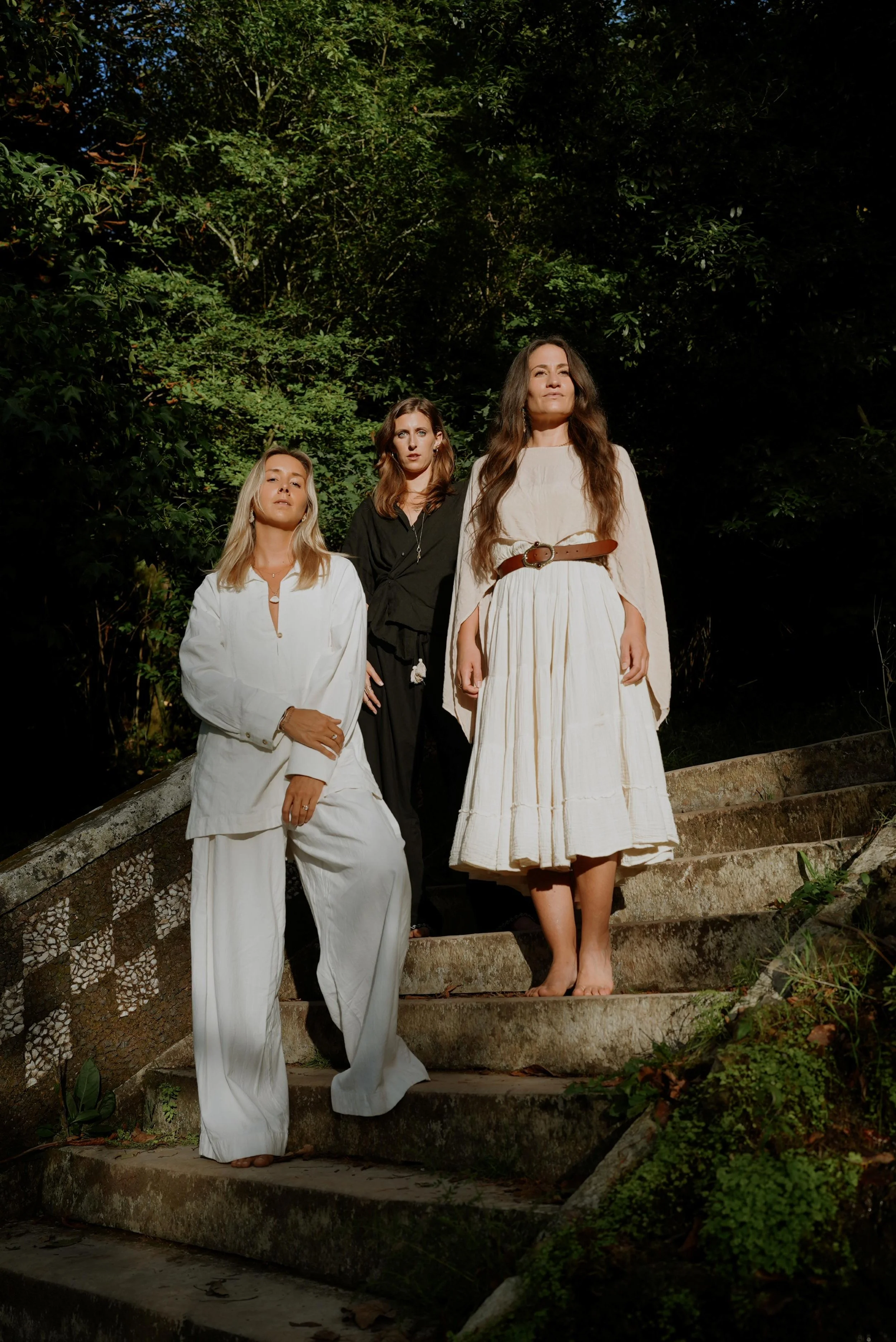 Three women standing on stone stairs outdoors surrounded by green foliage, dressed in white, black, and beige clothing, with serious expressions.