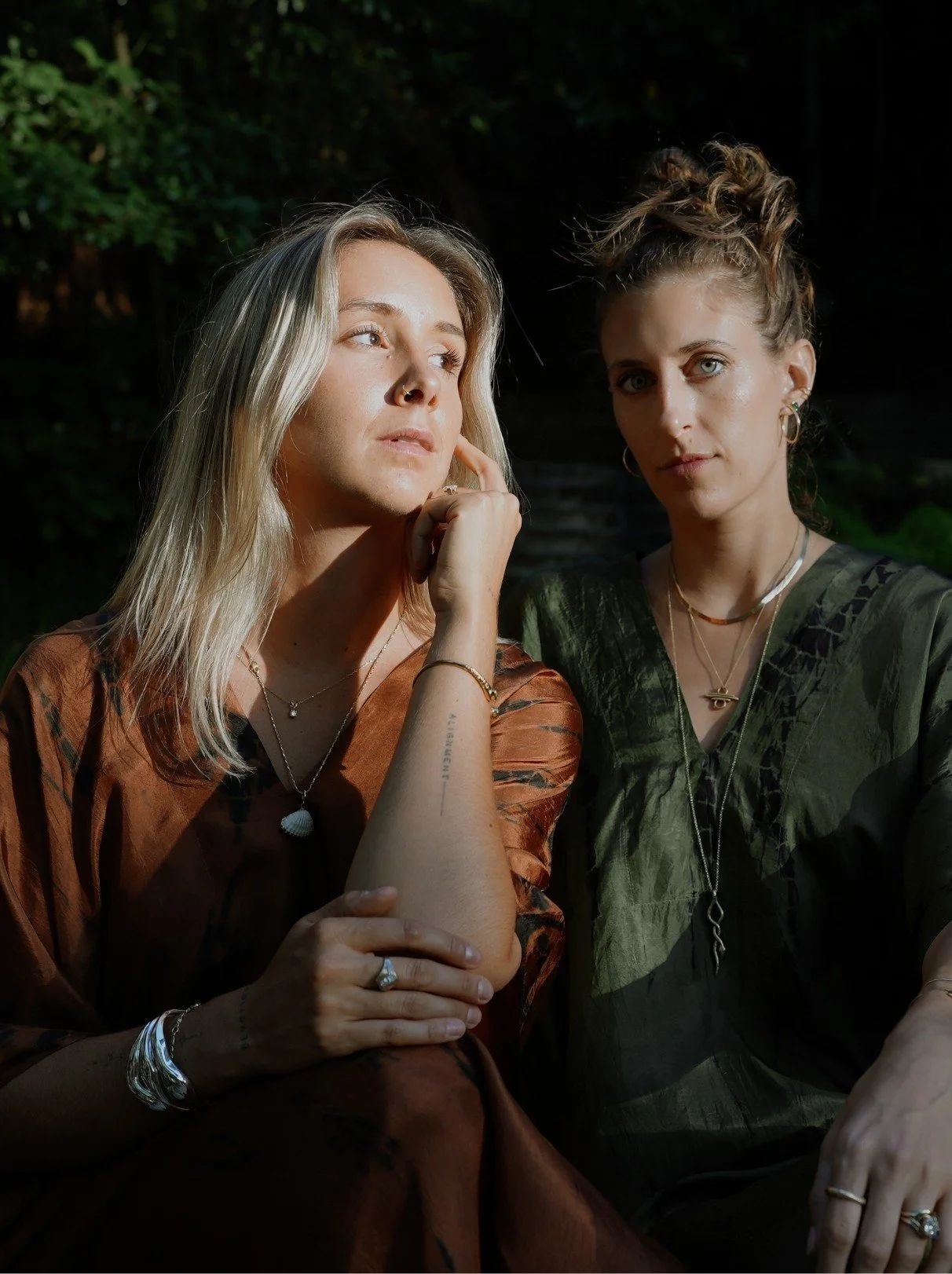 Two women outdoors at night, one with blonde hair and the other with curly light brown hair, both wearing jewelry, looking directly at the camera. The blonde woman rests her chin on her hand, wearing a rust-colored top, while the other woman wears a green top.