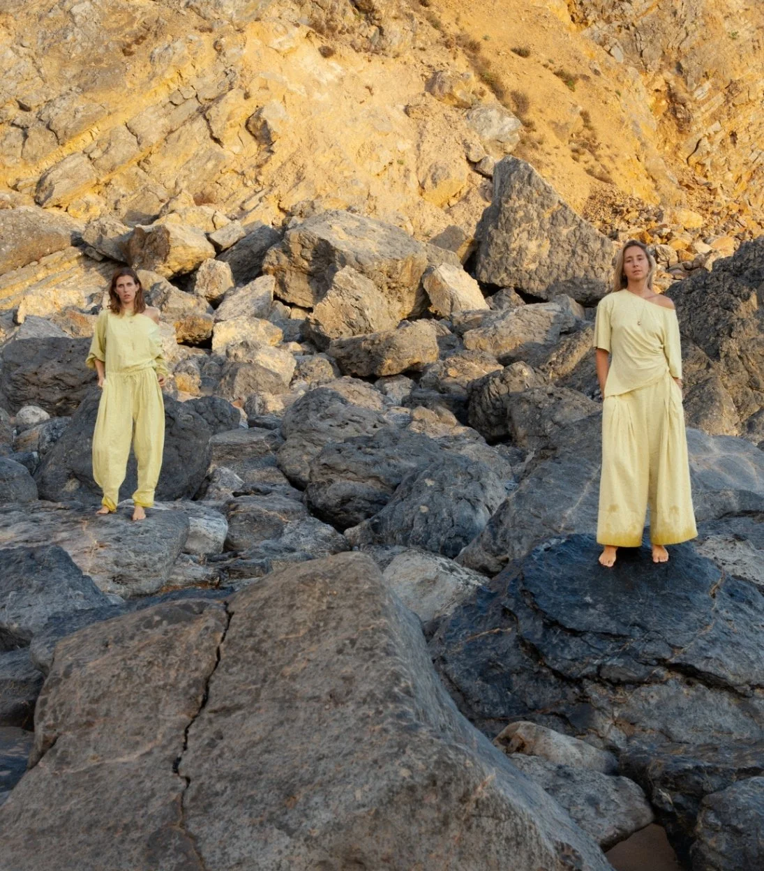 Two women in yellow outfits standing on rocks in a rocky terrain with a cliff in the background.