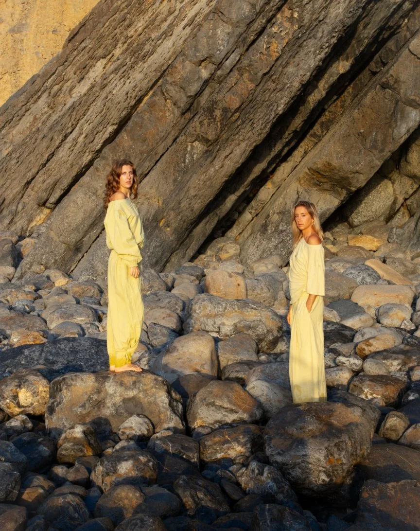 Two women in yellow dresses standing on large rocks in front of a steep rock face.