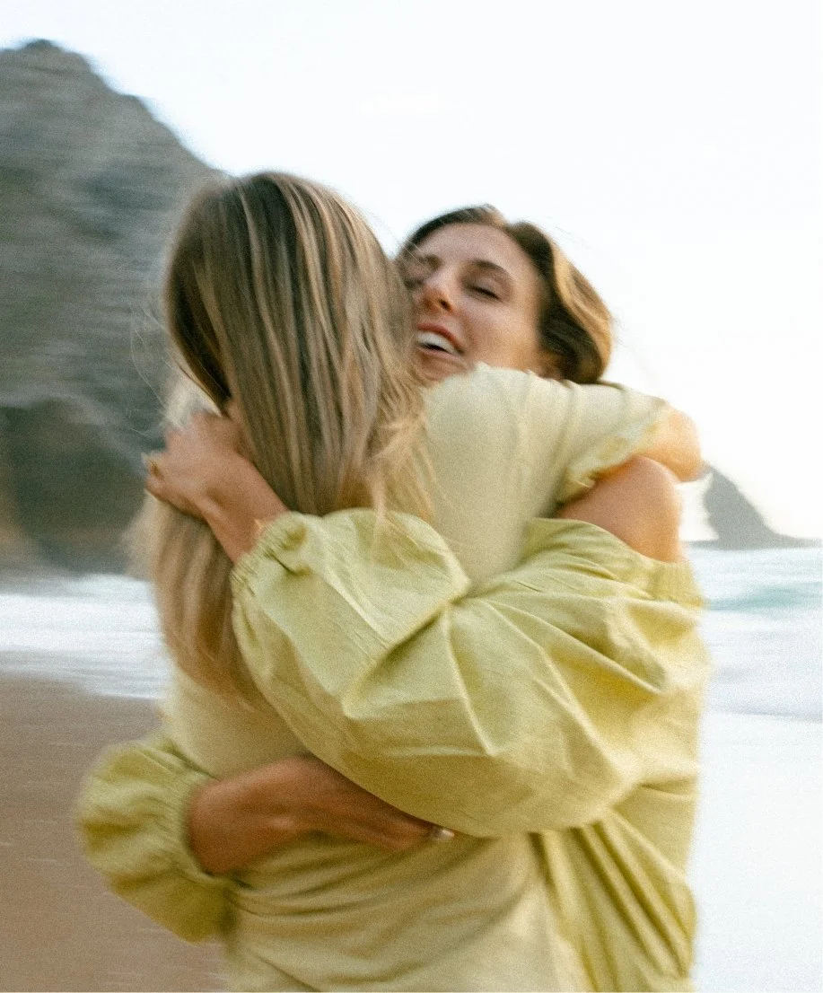 Two women hugging and smiling at the beach with cliffs in the background.