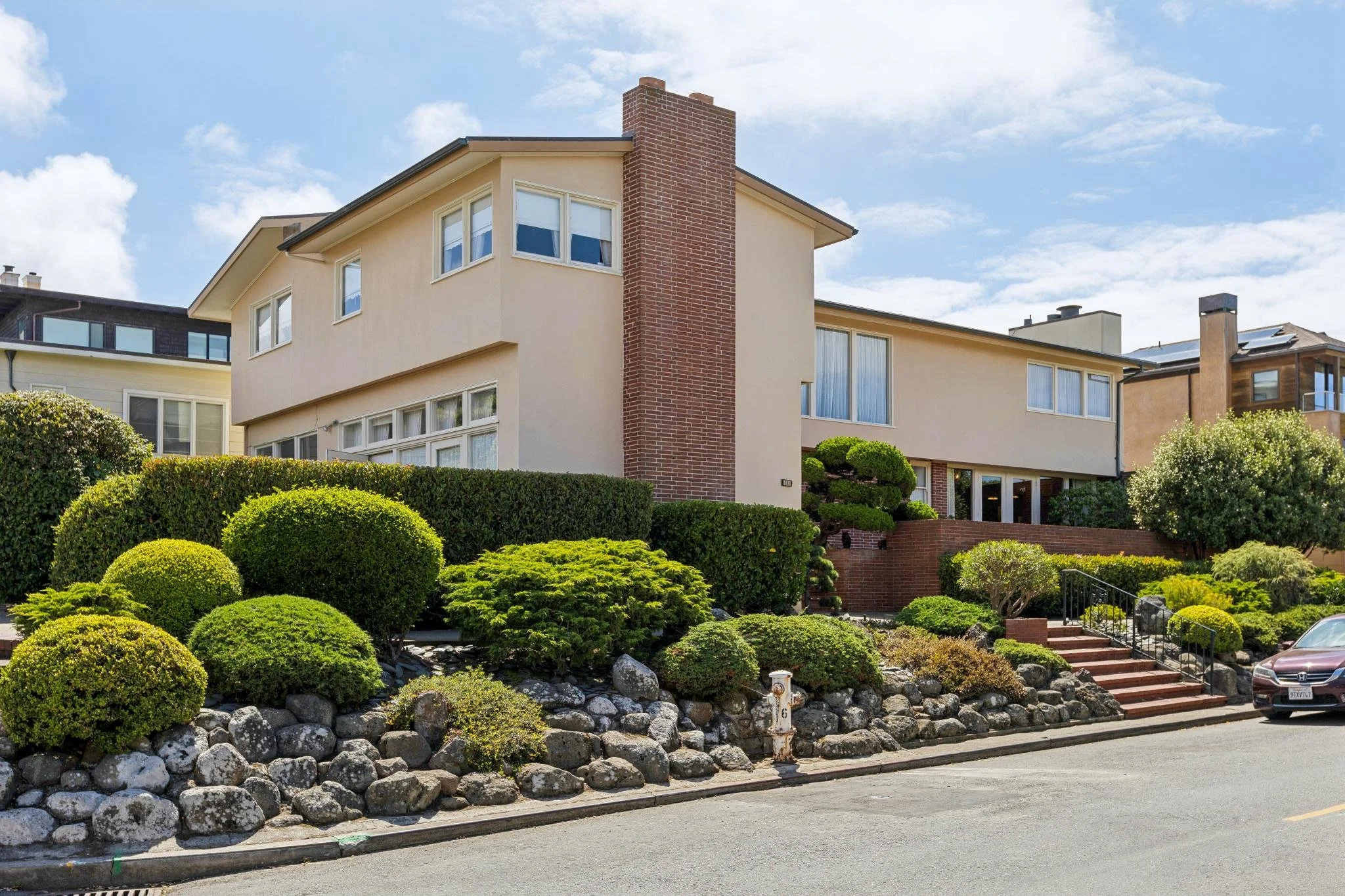 A multi-story house with beige stucco walls, a prominent brick chimney, and large windows. In front, there is a landscaped yard with various green bushes and rocks along the slope, and a street with parked cars.