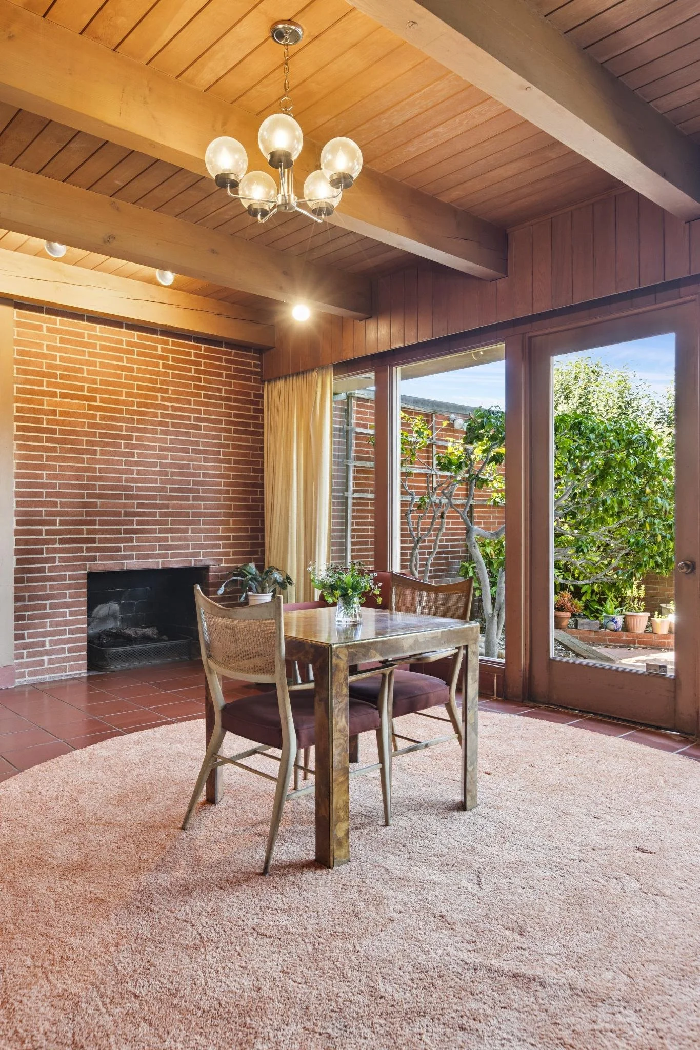 Interior of a room with wood-paneled ceiling, brick fireplace, glass doors, and a dining table with four chairs, decorated with potted plants and visible outside greenery.