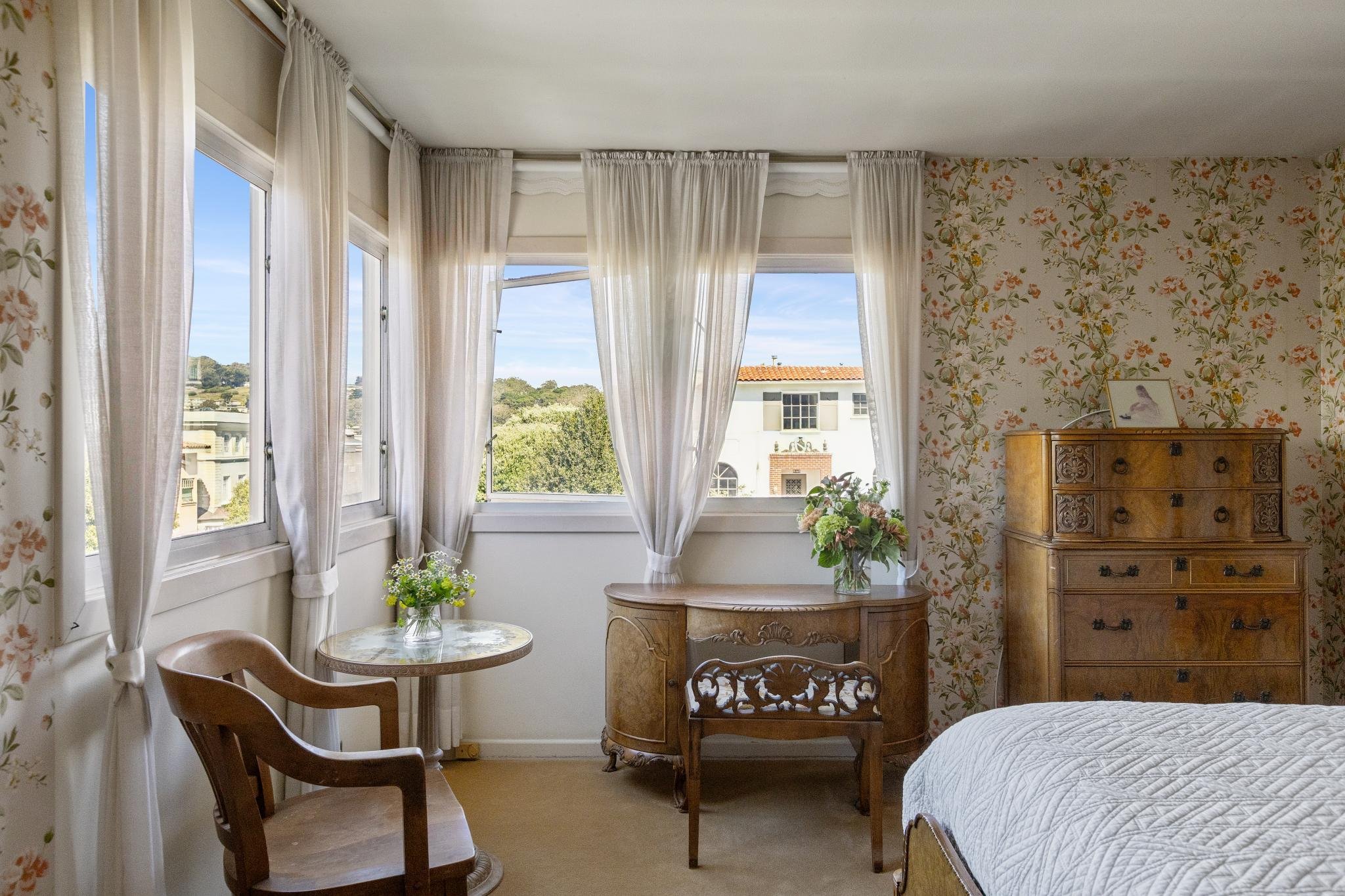 A cozy bedroom with three large windows covered with white curtains, overlooking a building and trees outside. The wall has floral wallpaper, and there is a vintage wooden dresser and a side table with a flower vase. Part of a bed with a white quilt 