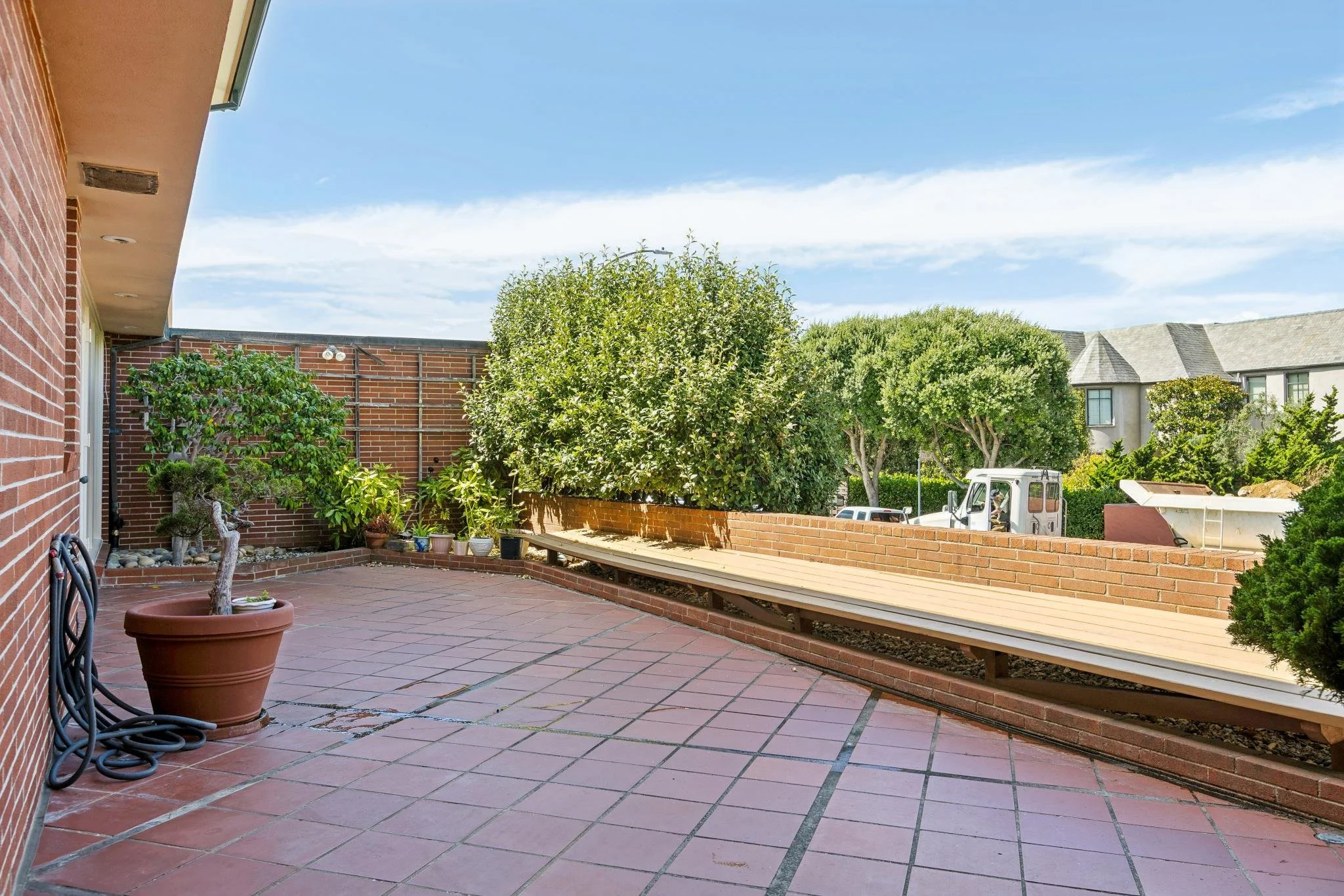 A spacious brick-paved backyard patio with potted plants, trees, and a wooden bench. The patio is enclosed by a brick wall, and there are houses and a blue sky with clouds in the background.