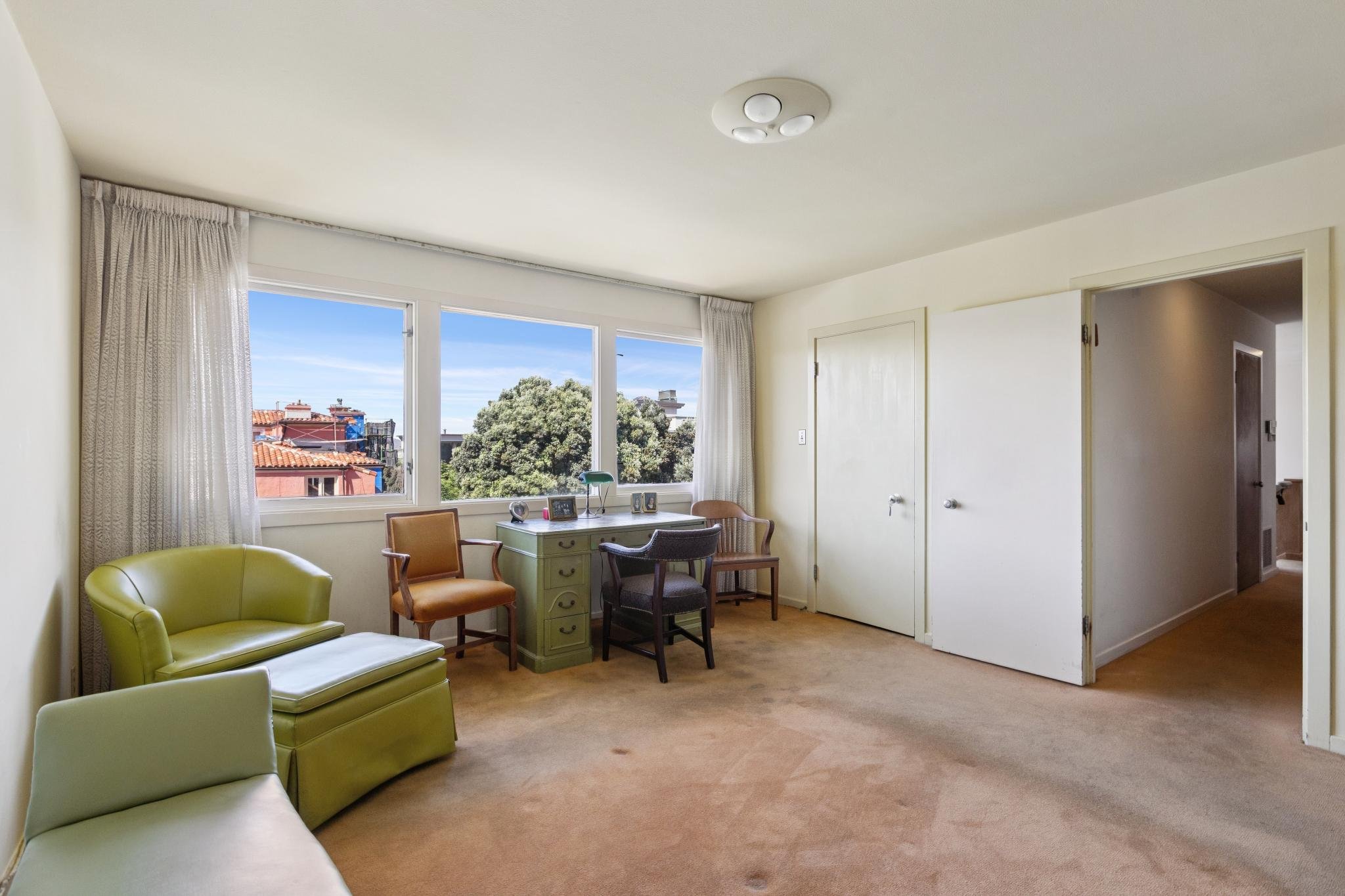 Living room with large windows showing rooftops and trees outside, featuring green and beige chairs, a small green desk, and a door on the right.