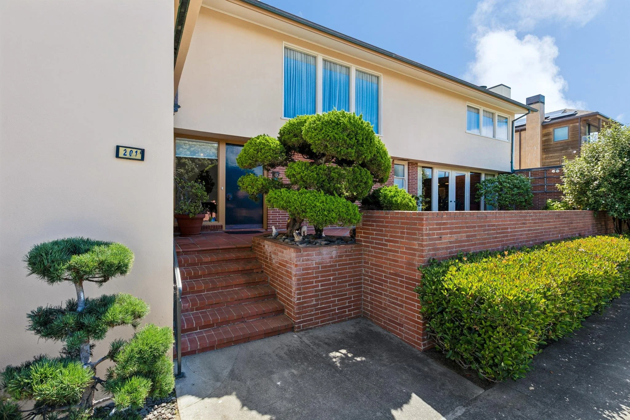 Front view of a modern townhouse with brick steps leading to the front door, surrounded by trimmed bushes and a small decorative tree, with a clear blue sky overhead.