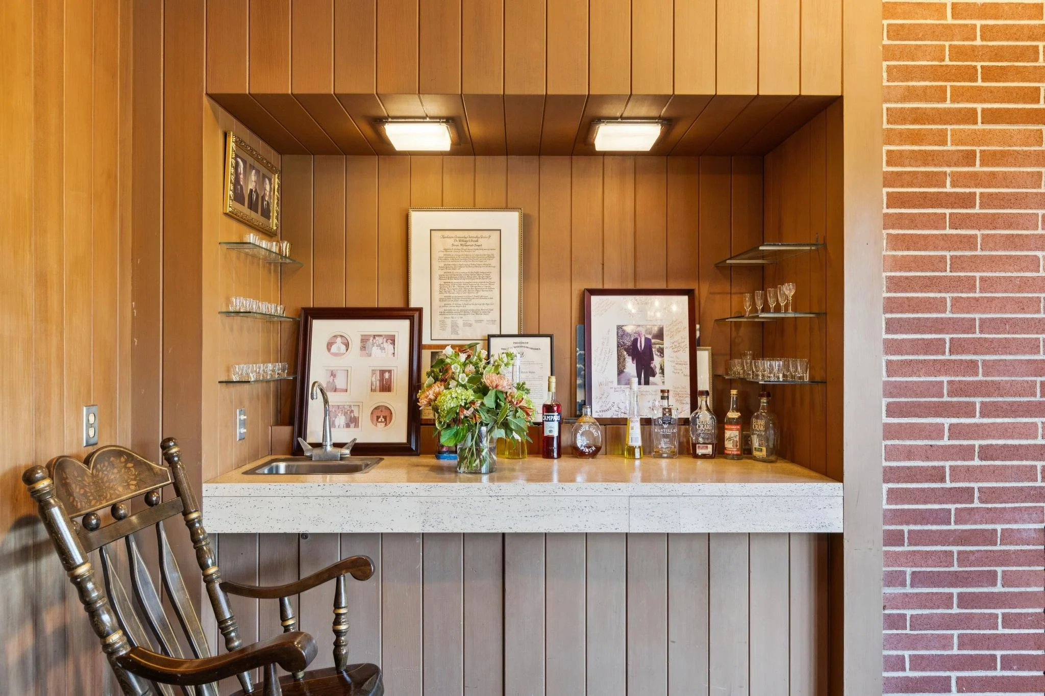 A kitchen counter with a flower arrangement, bottles, framed photographs, family pictures, and wall art, with wooden paneling on the wall and a brick column on the right.