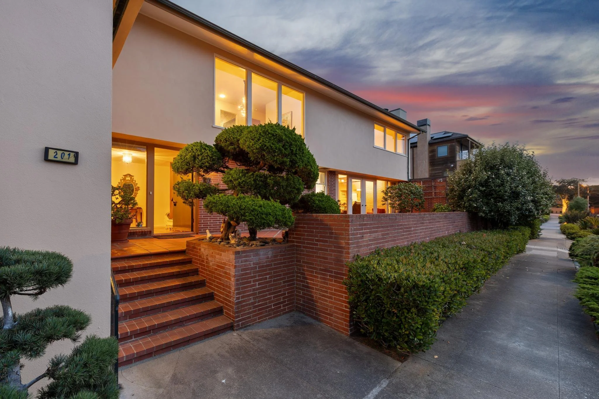 Front view of a modern house during sunset with large windows, brick steps, and lush green shrubbery.