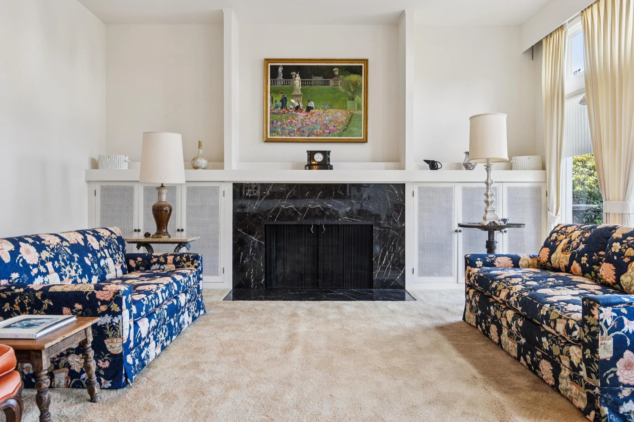 A living room with two floral-patterned sofas facing each other, a beige carpet, a fireplace with black marble, and a white wooden mantle. Above the fireplace, there is a framed landscape painting of a garden scene. On the mantle, there are decorativ
